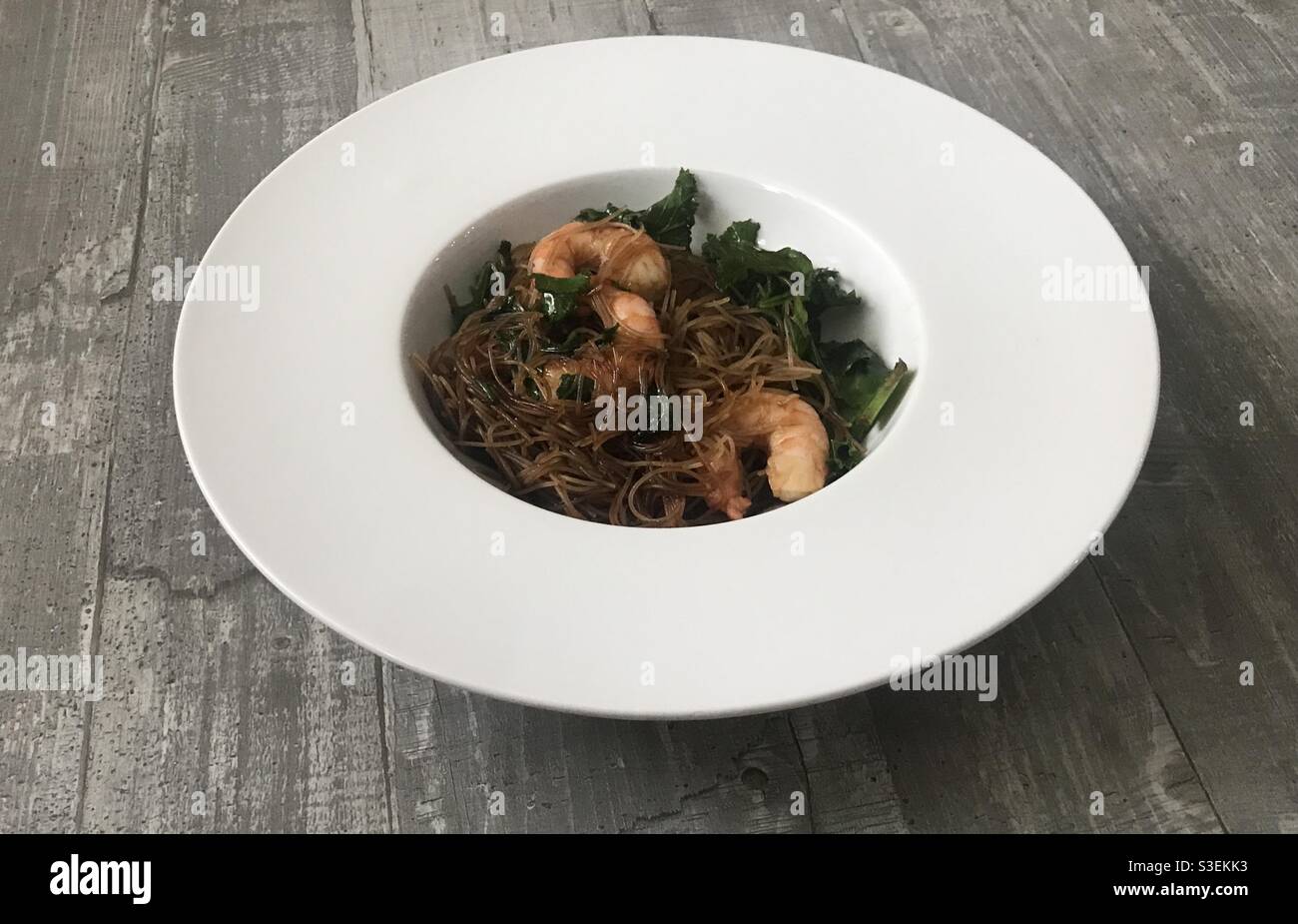 Closeup image of a bowl of honey sesame prawns served with rice noodles against a wooden background - Smartphone Captured Stock Image