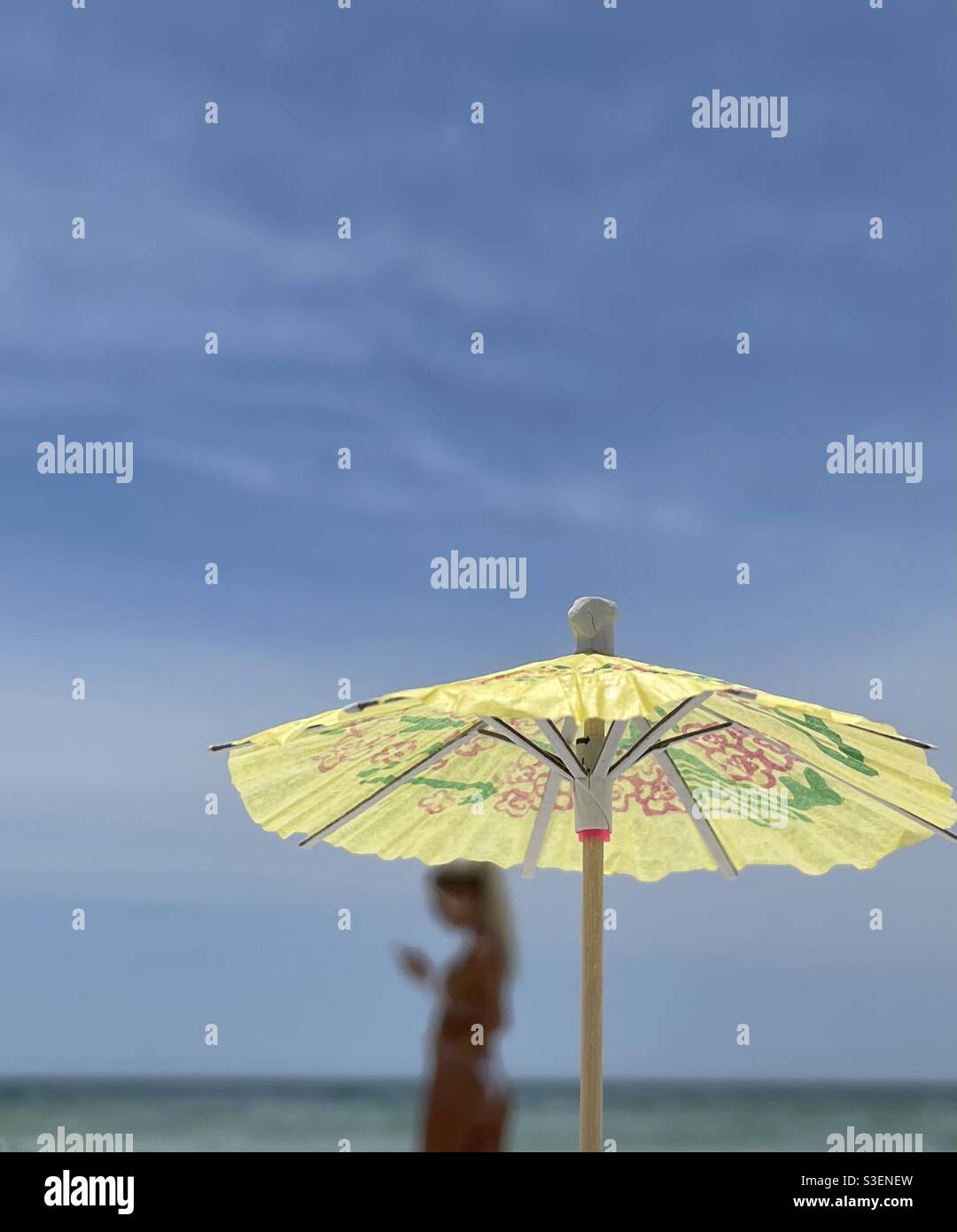 Select focus on miniature colorful umbrella with blurred woman and ocean water background - Smartphone Captured Stock Image