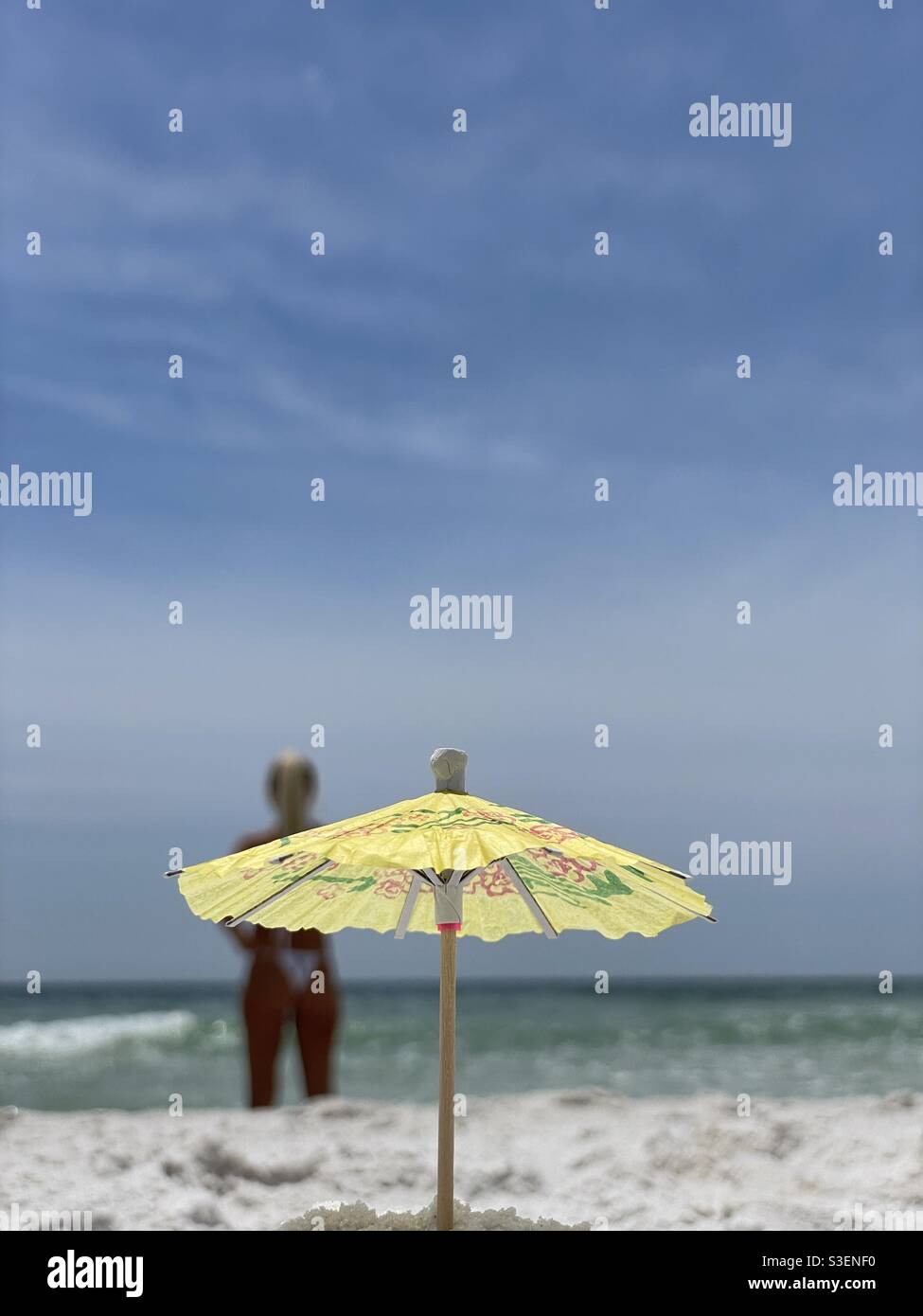 Select focus on miniature umbrella with blurred woman and beach background - Smartphone Captured Stock Image
