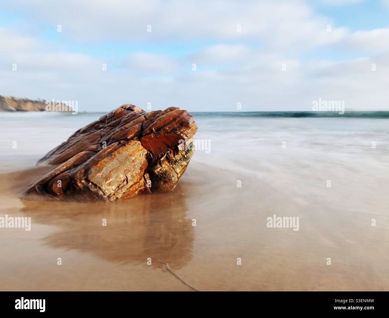 Long exposure shot of rock on sandy beach with ocean water washing up. Stock Photo