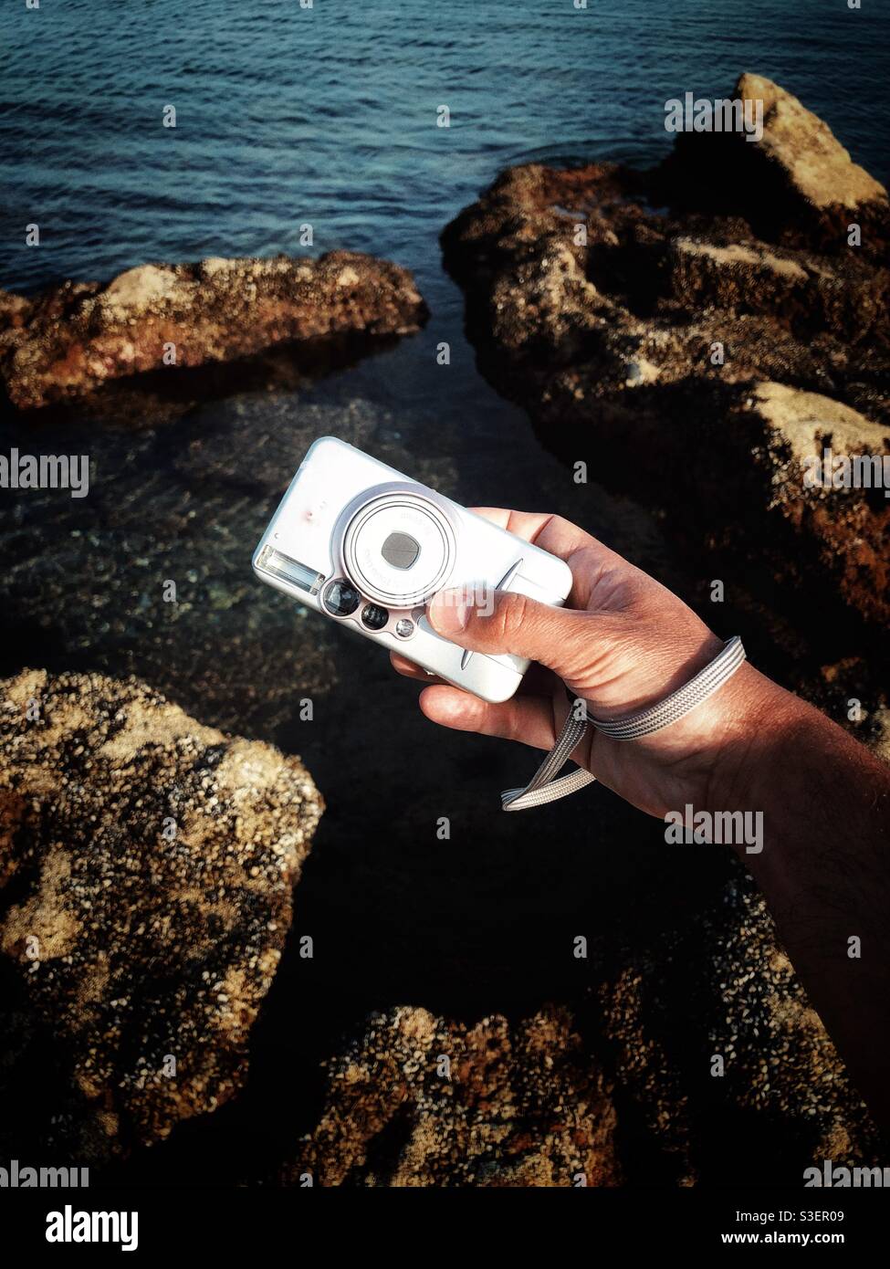 Man holding a point and shoot camera over the rocky shore - Smartphone Captured Stock Image