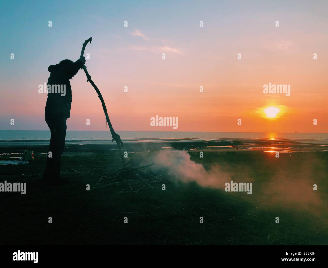 A man builds a bonfire in front of a sunset over the sea. Camping holiday. Walney island. Cumbria. Uk - Smartphone Captured Stock Image