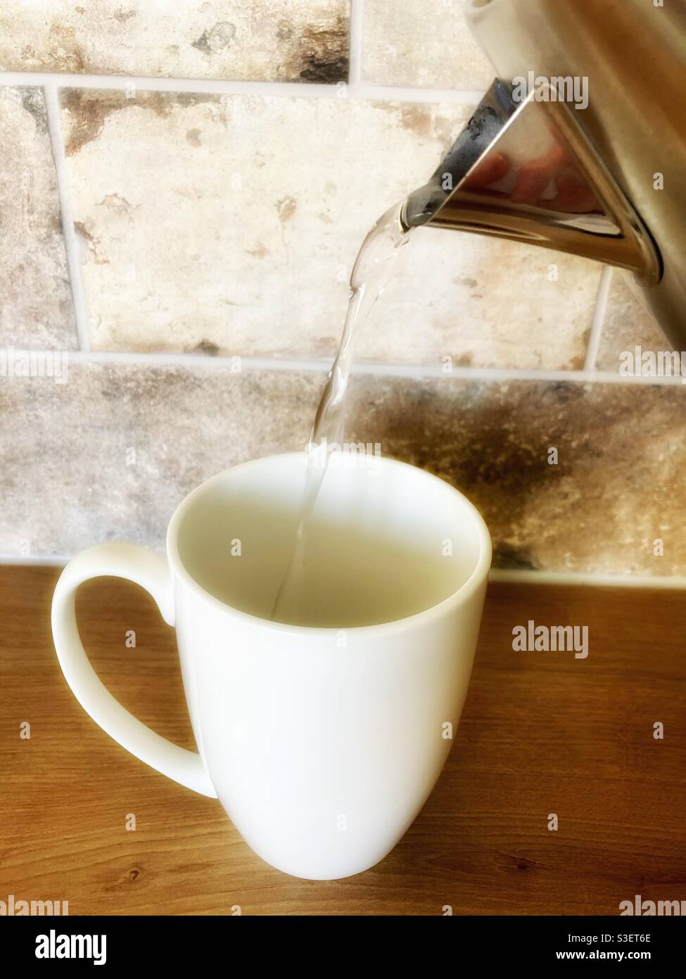 Pouring hot water from a kettle into a white mug to make a cup of tea in the kitchen Stock Photo