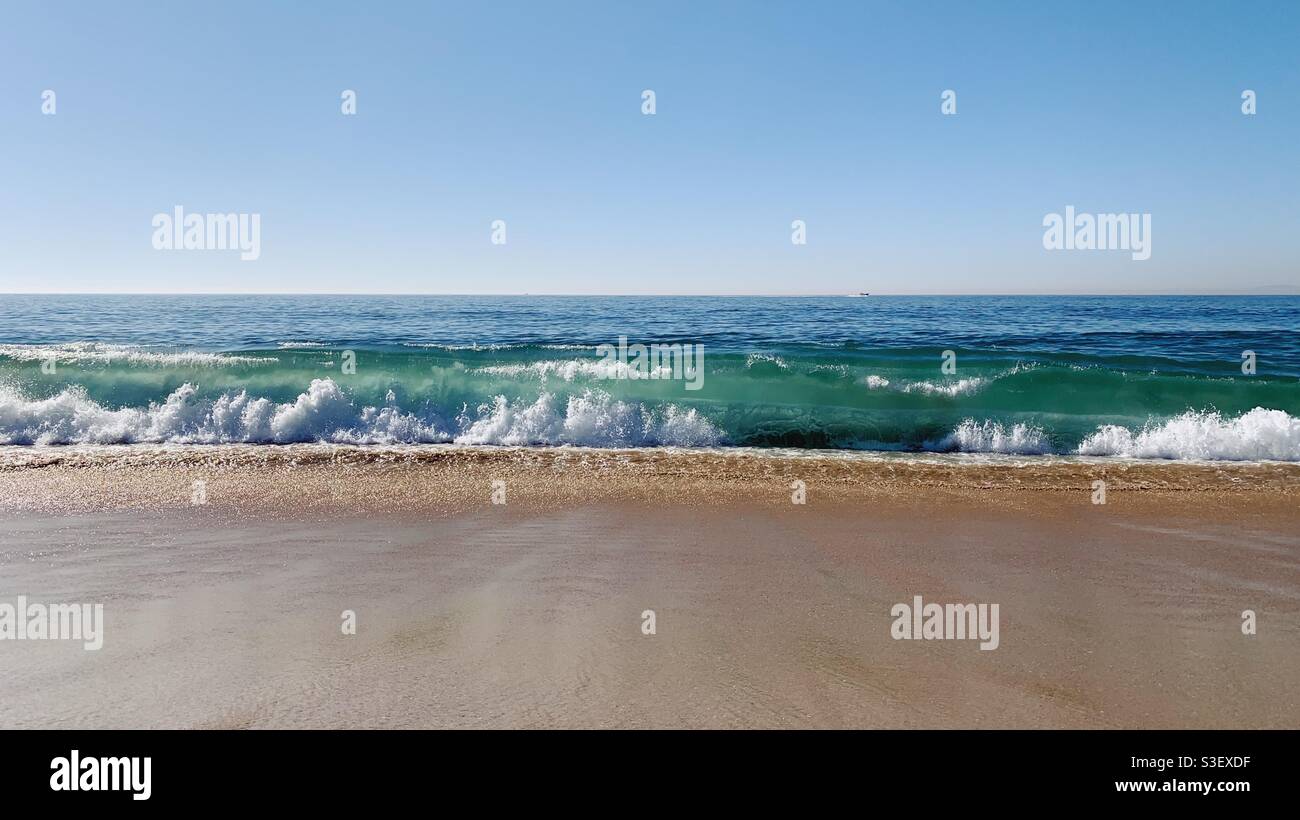 Ocean wave breaking at sandy beach under a clear blue sky. - Smartphone Captured Stock Image