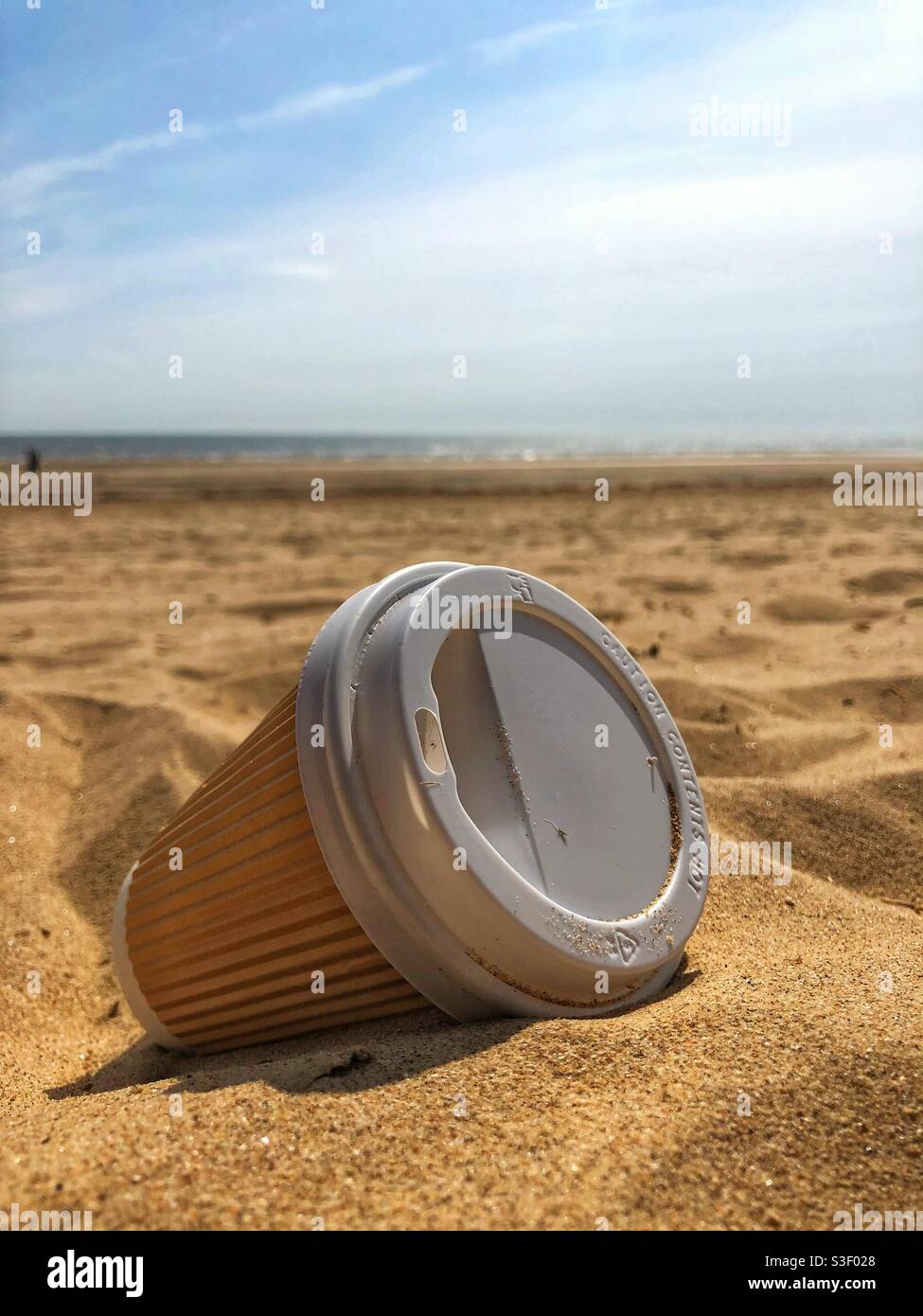 A single use takeaway non-recyclable cardboard and plastic coffee cup washed up on a sandy beach in an environmental issues concept - Smartphone Captured Stock Image