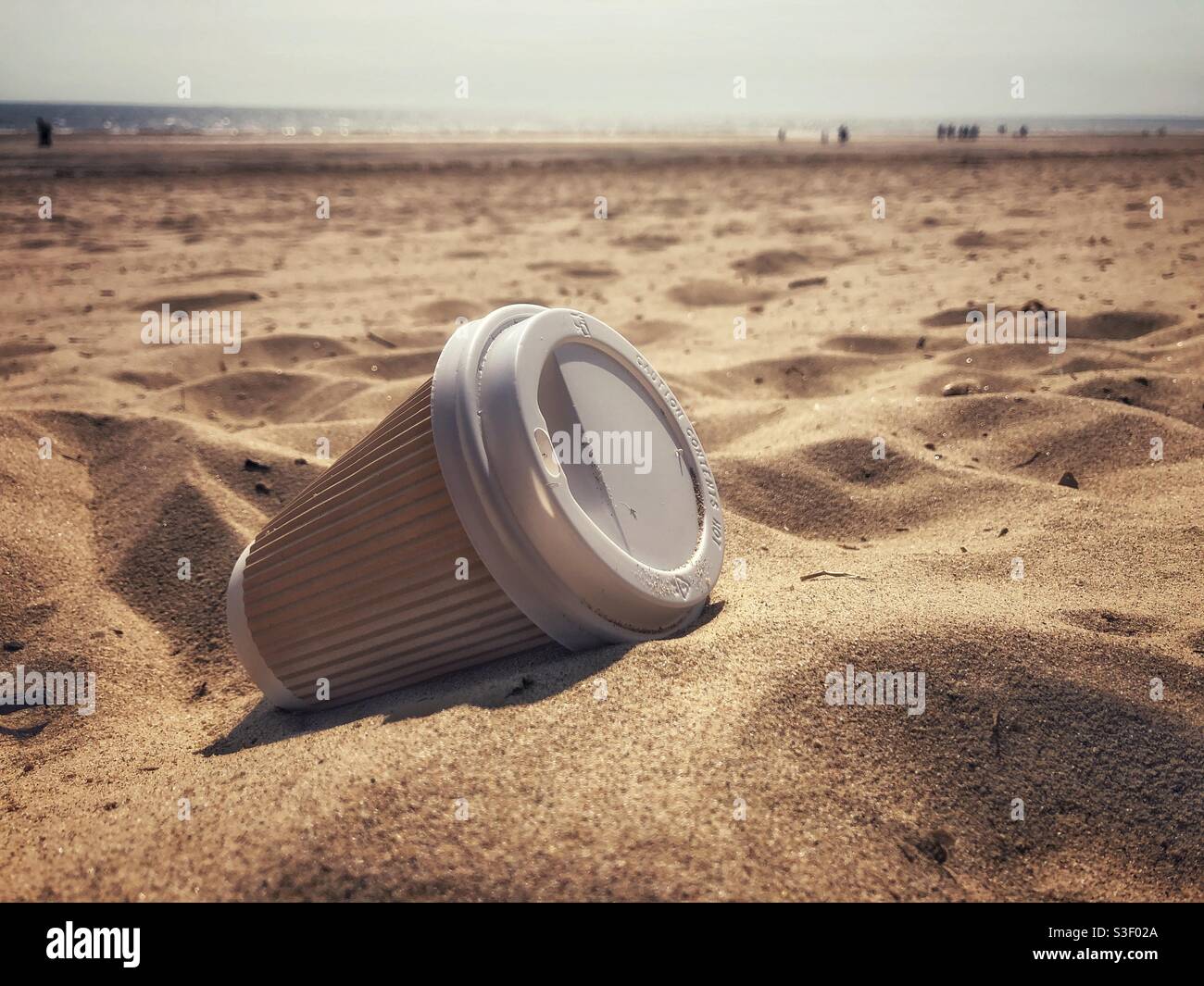 A discarded cardboard and plastic single use takeaway coffee cup lying on a beautiful sandy beach in an environmental issues concept - Smartphone Captured Stock Image