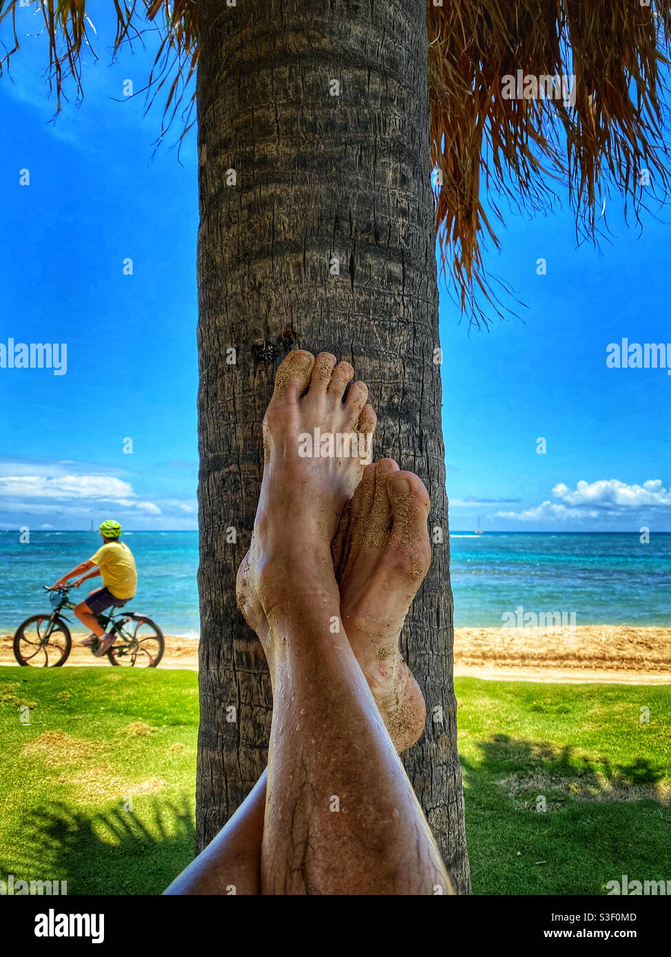 Sandy feet resting against the trunk of a palm tree as a bicyclist pedals past Stock Photo