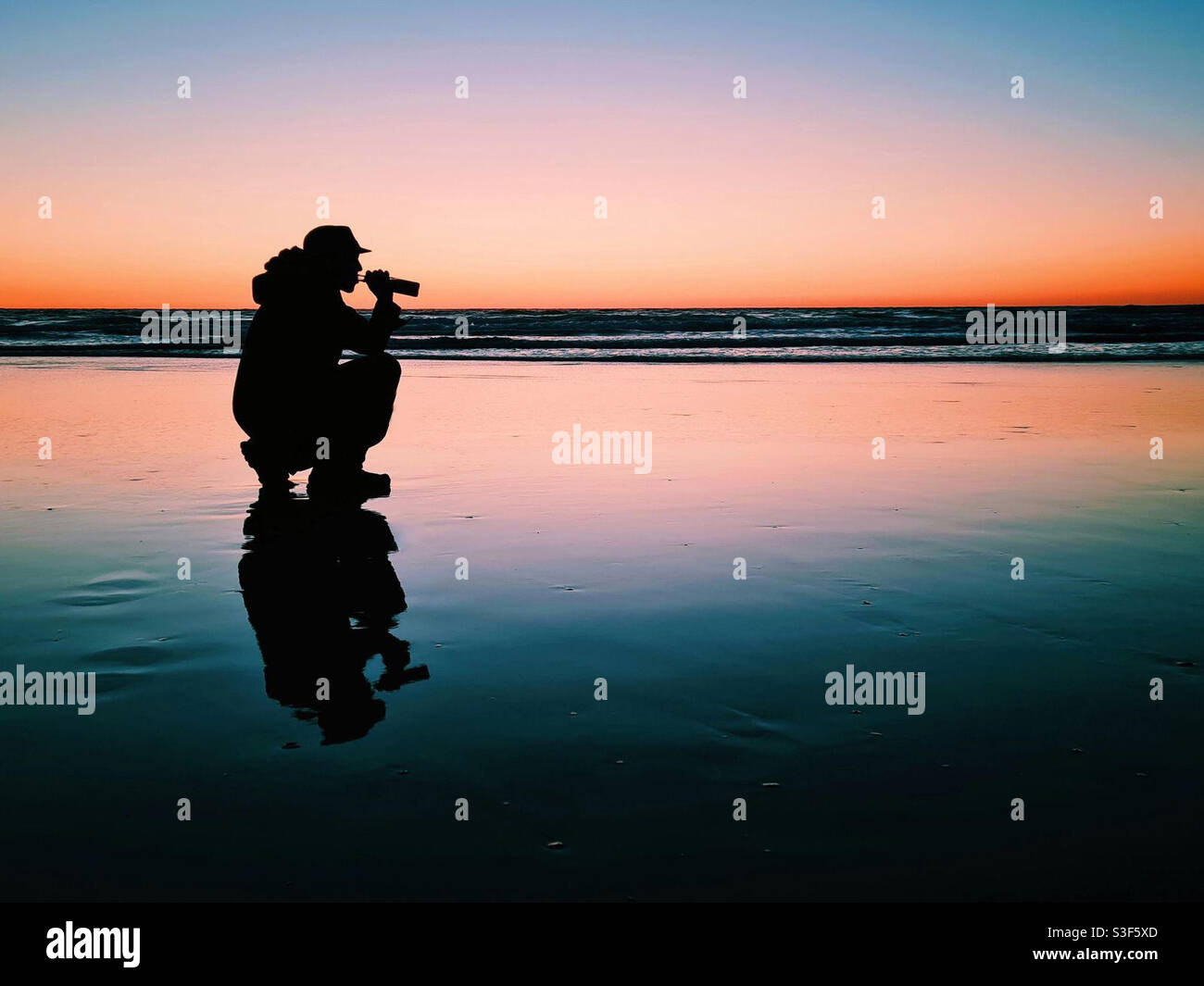 A silhouette of a man drinking from a bottle of beer on the beach at sunset in Cornwall. Travel photography. Staycation England, Uk - Smartphone Captured Stock Image