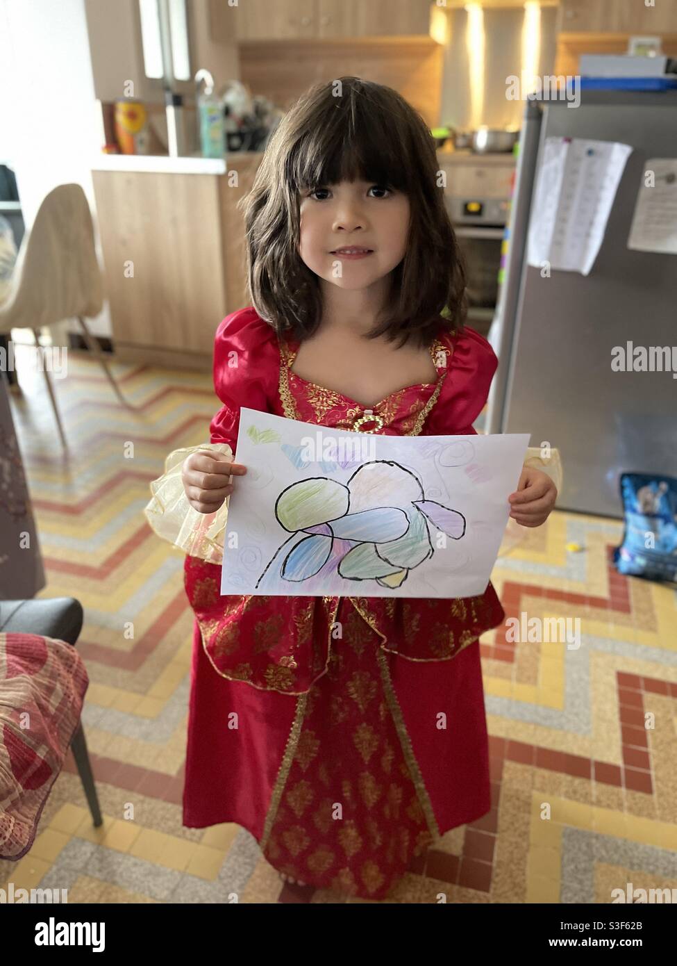 Beautiful and cute mixed Asian little girl with long brown hair wearing a red dress with gold trim holding a nice colour drawing, smiling Stock Photo