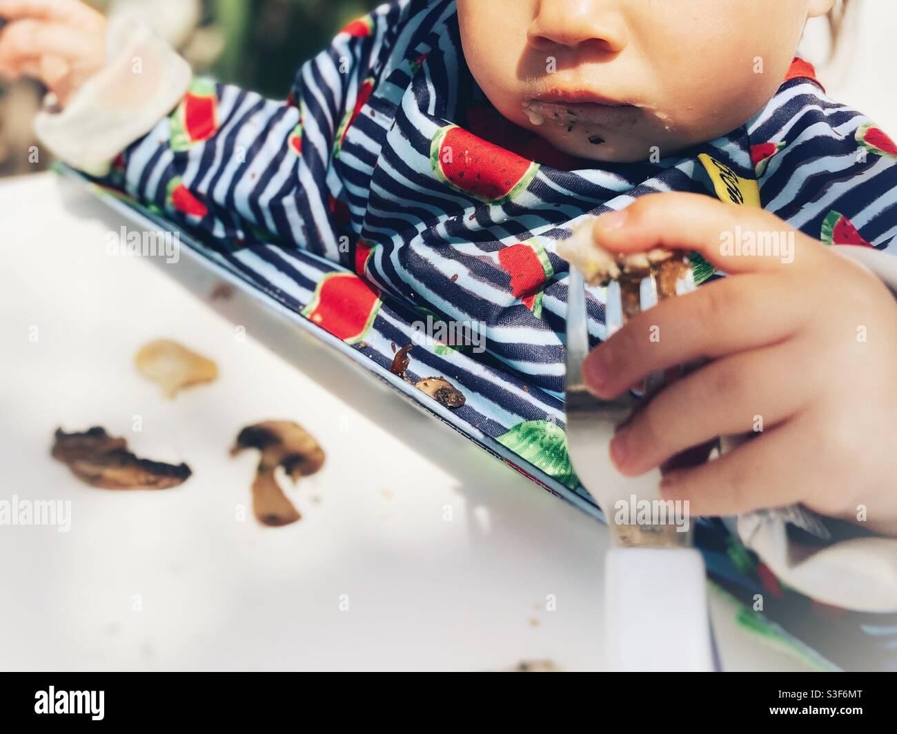 Baby/ infant/ toddler/ child eating cooked mushrooms in a high chair holding a child’s fork Stock Photo