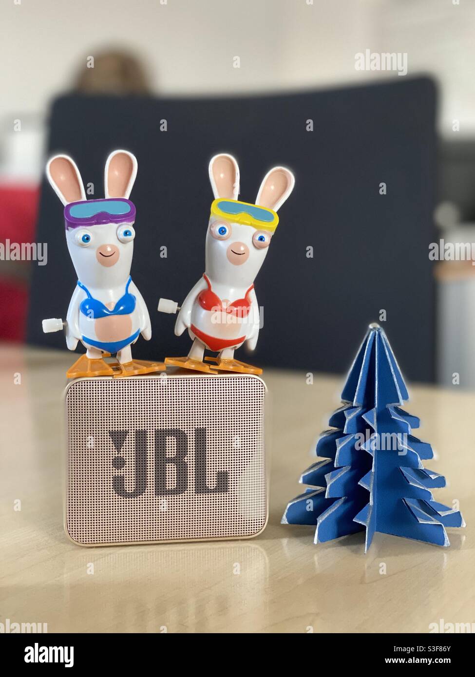 Two toy Raving Rabbits standing on top of a golden JBL Go Bluetooth speaker, with a green paper Christmas tree on an office desk - Smartphone Captured Stock Image