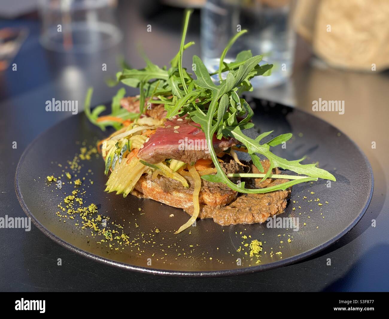 Flank steak atop Fois Gras and meat sauce covered by Arugula leaves on a black restaurant plate in Vandargues, near Baillargues and Montpellier, Occitanie, south of France - Smartphone Captured Stock Image