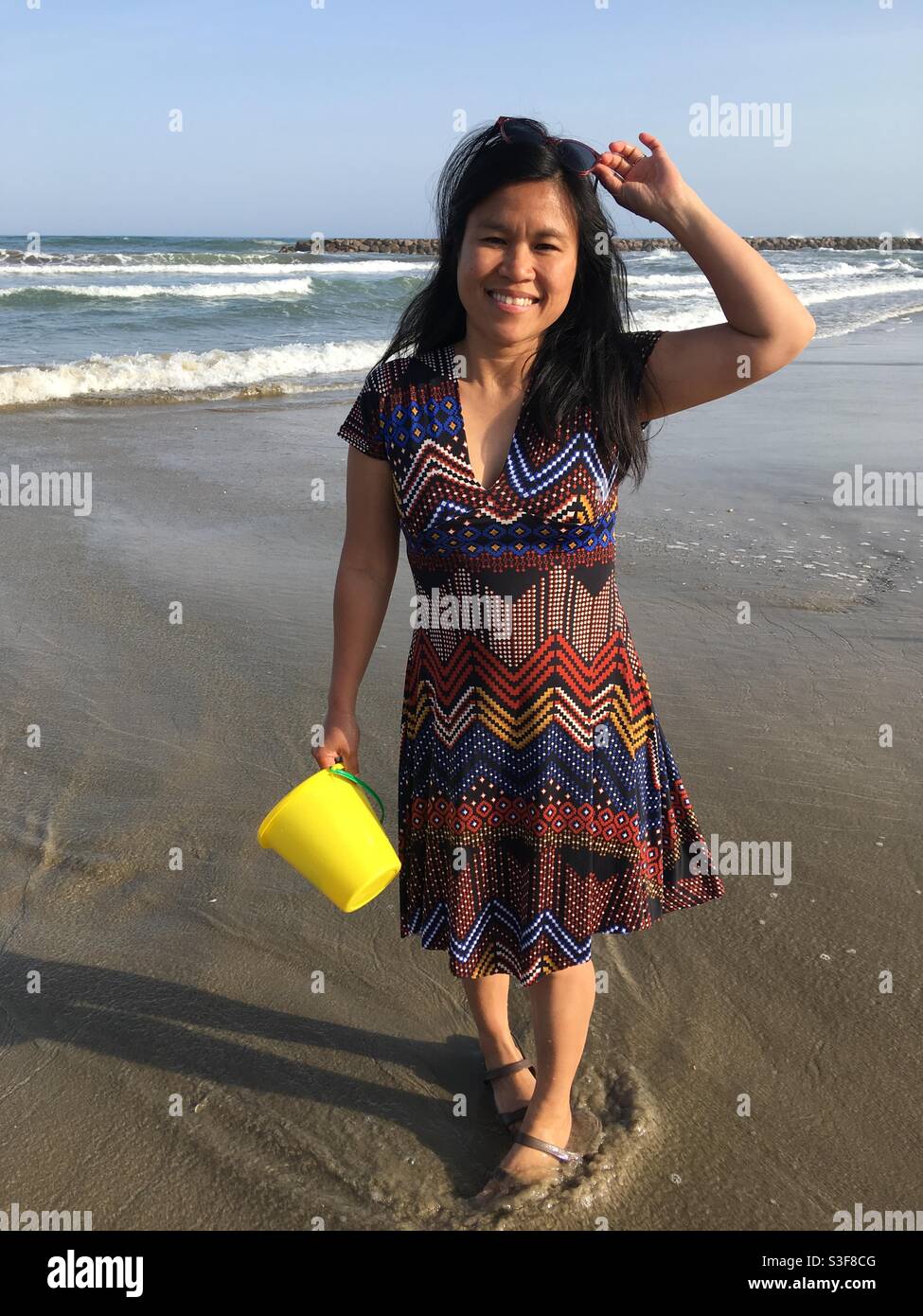 Cute Asian Filipino girl standing on the beach near the water with a yellow beach toy wearing a colorful pattern dress in Palavas les Flots near Carnon Plage, Montpellier, Occitanie, south of France Stock Photo