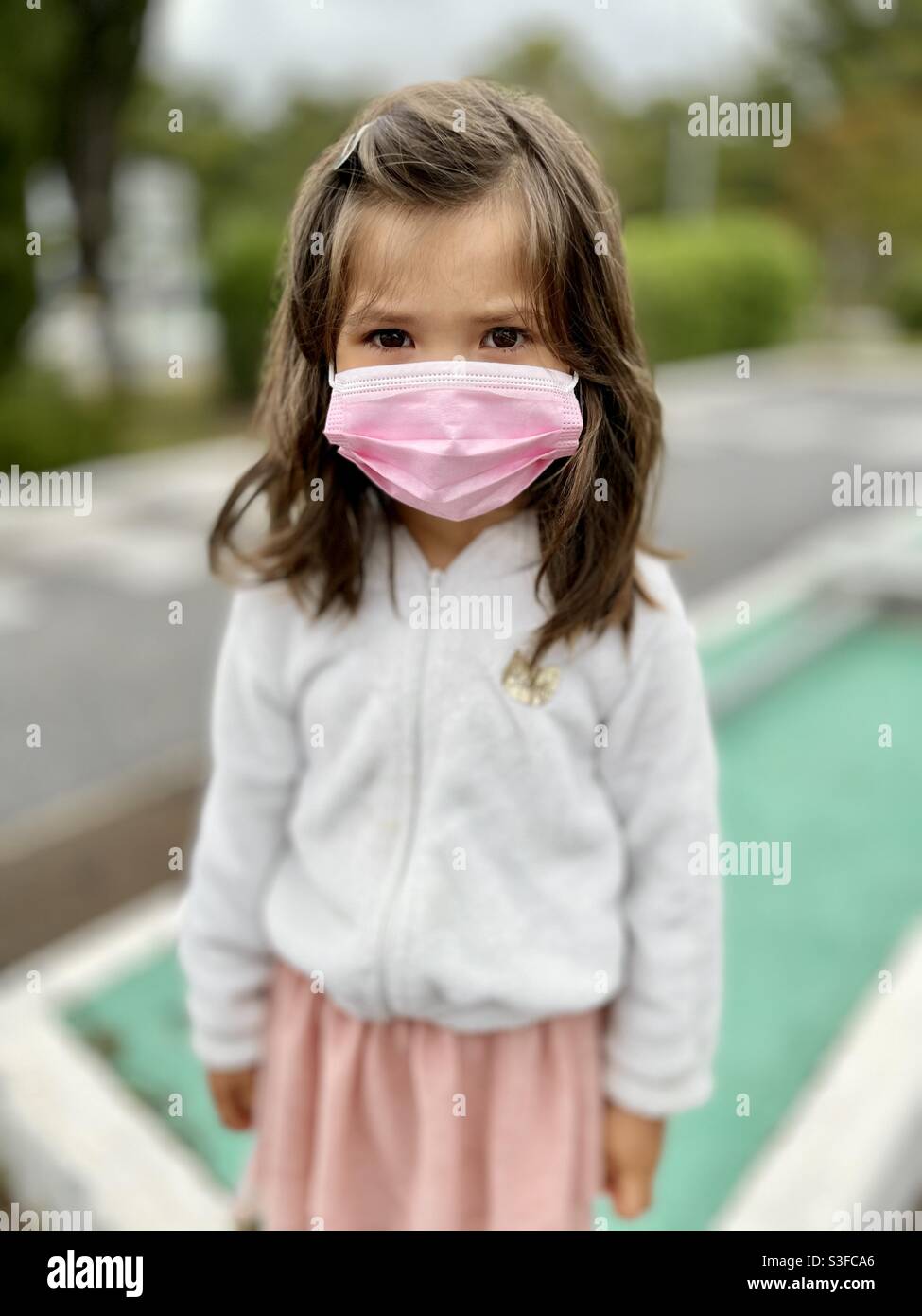 Mixed Asian Filipino little girl with long brown hair wearing a pink mask and a white fur hoodie and pink dress on a blurred background Stock Photo