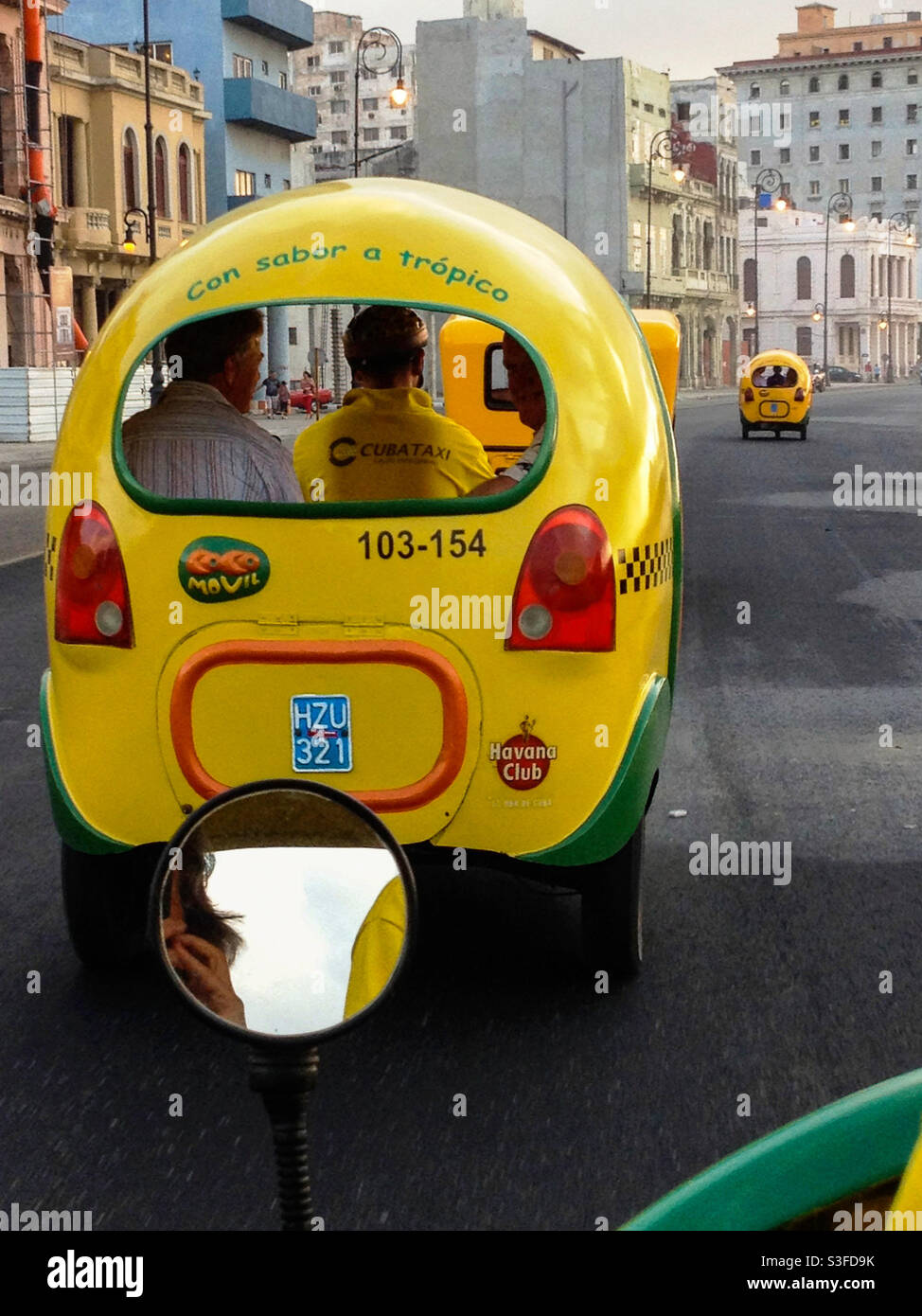 View from behind of a quirky cute yellow Coco taxi with driver and tourist passengers in Havana, Cuba Stock Photo