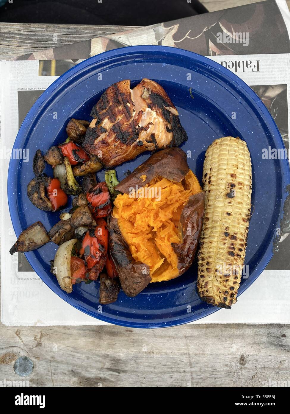 A delicious meal of salmon, corn on the cob, sweet potato and mixed vegetables grilled on an open fire served on a blue enameled tin plate. - Smartphone Captured Stock Image