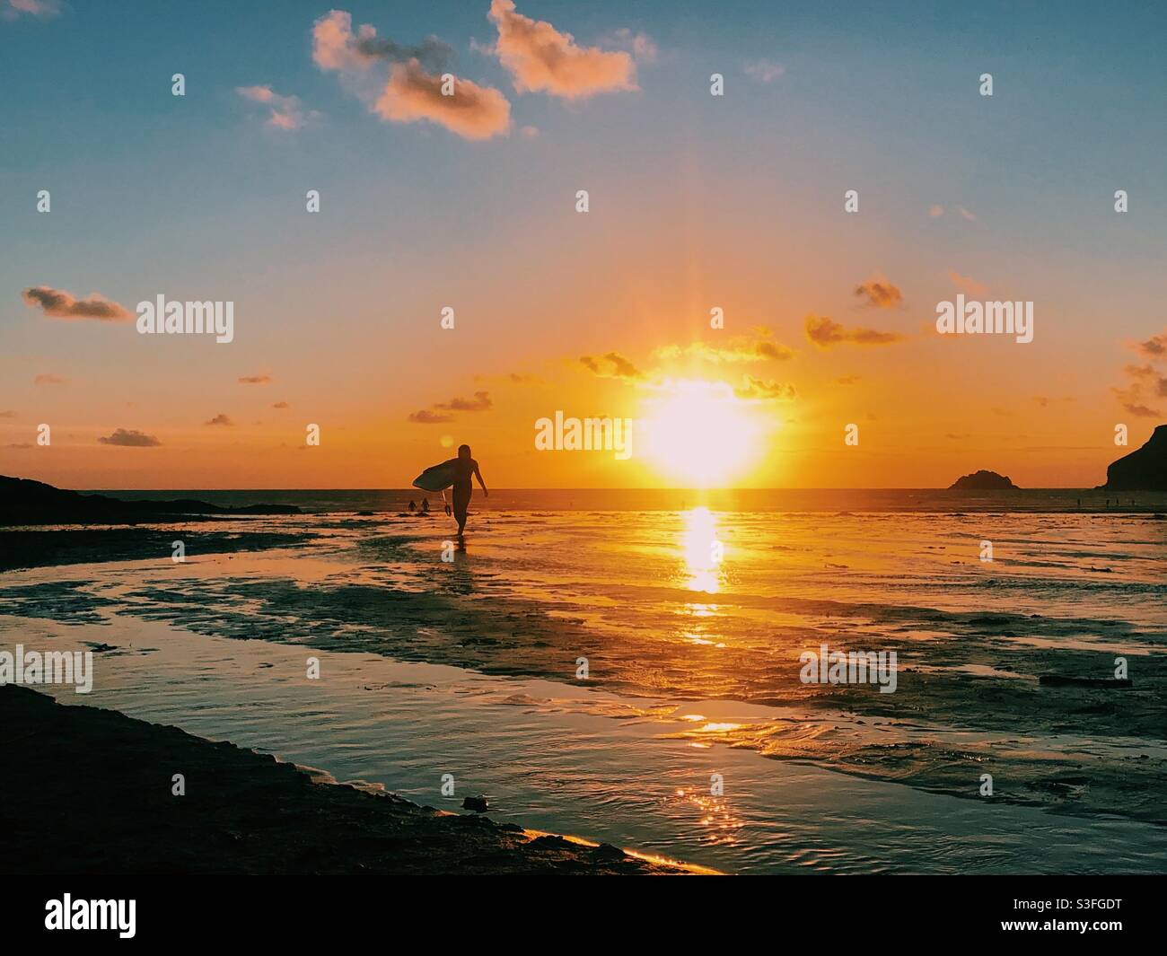 A surfer walks along the beach silhouetted in front of a beautiful sunset in Cornwall, England - Smartphone Captured Stock Image