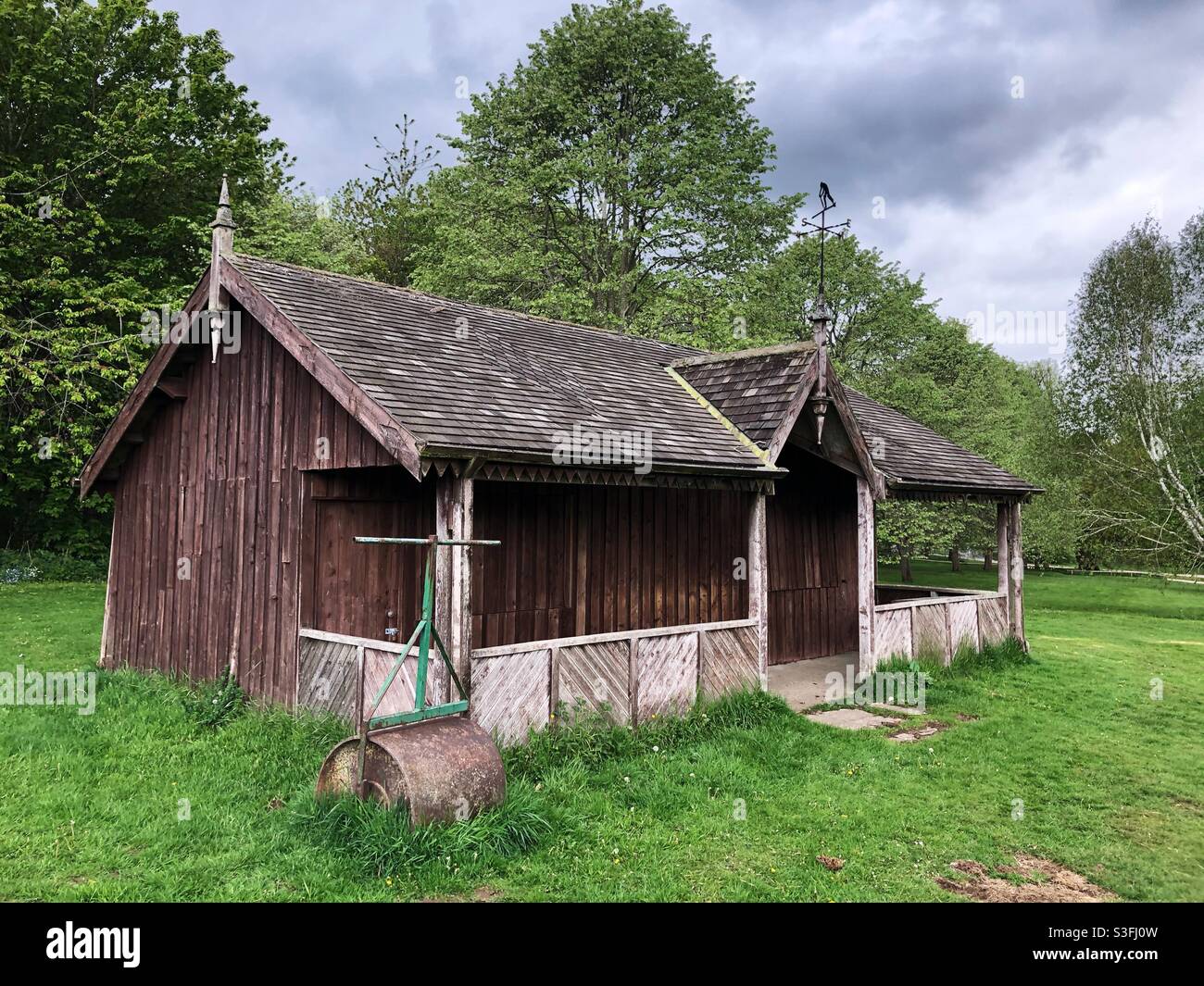 An old wooden cricket pavilion with lawn roller in a traditional English countryside village setting Stock Photo