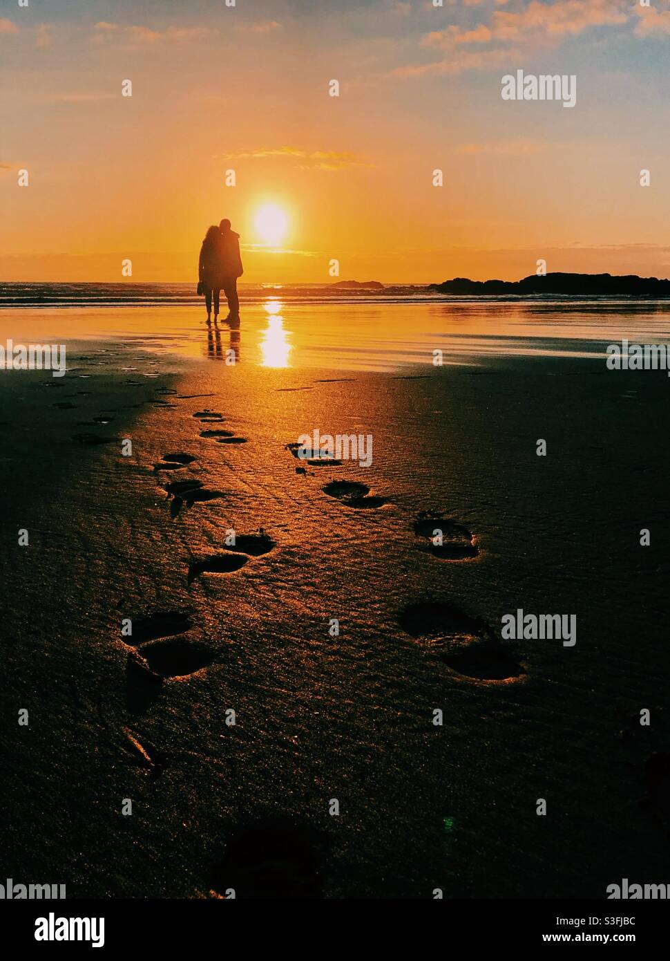 A couple silhouetted watching the sunset on a beach, footprints leading to them Stock Photo