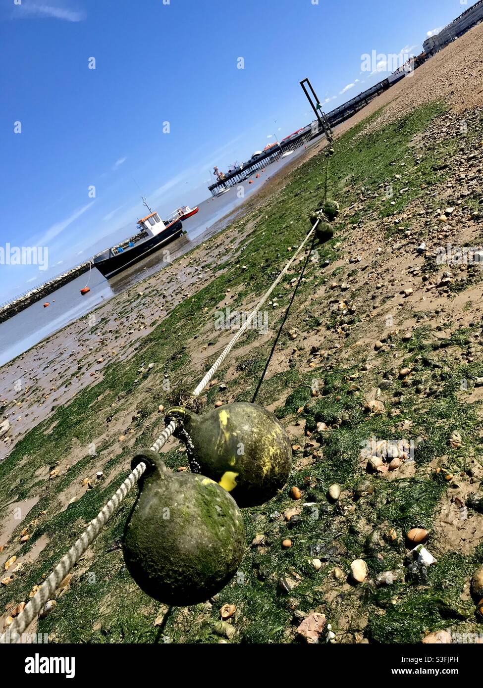 Buoys and ropes on a beach, at low tide, covered in algae with a fishing boat anchored in the background on a spring day, in Herne Bay, Kent, UK. - Smartphone Captured Stock Image