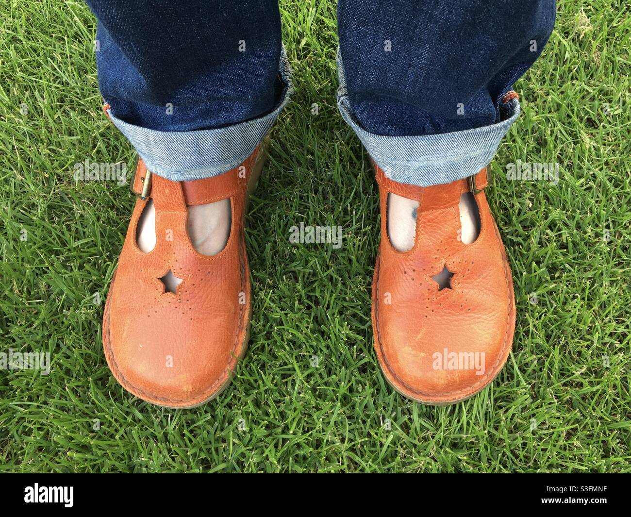 A woman’s feet in orange Mary Jane style flat shoes standing on green grass and wearing blue jeans Stock Photo