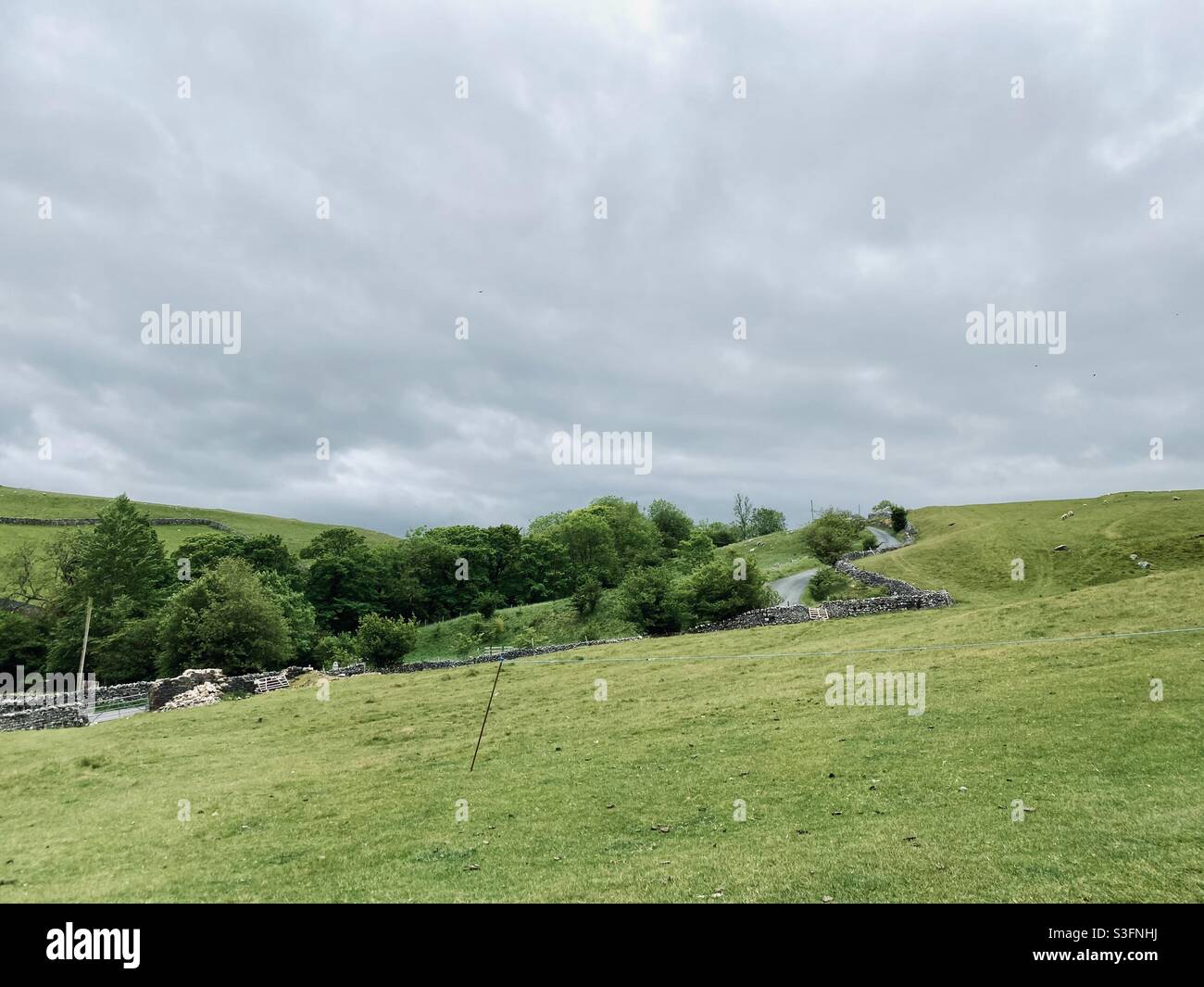 Scenic rural landscape with dramatic grey sky in Malham, Yorkshire Dales National Park, UK. Rolling hills and green pastures with dry stone walls and winding road - Smartphone Captured Stock Image
