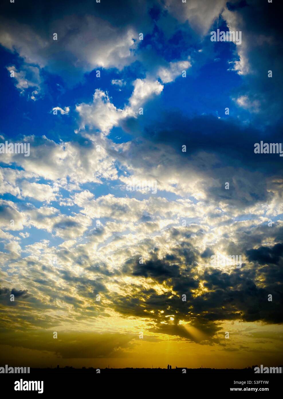 Bright golden sunset under a sky spangled with clouds, silhouetting a small city below. Stock Photo