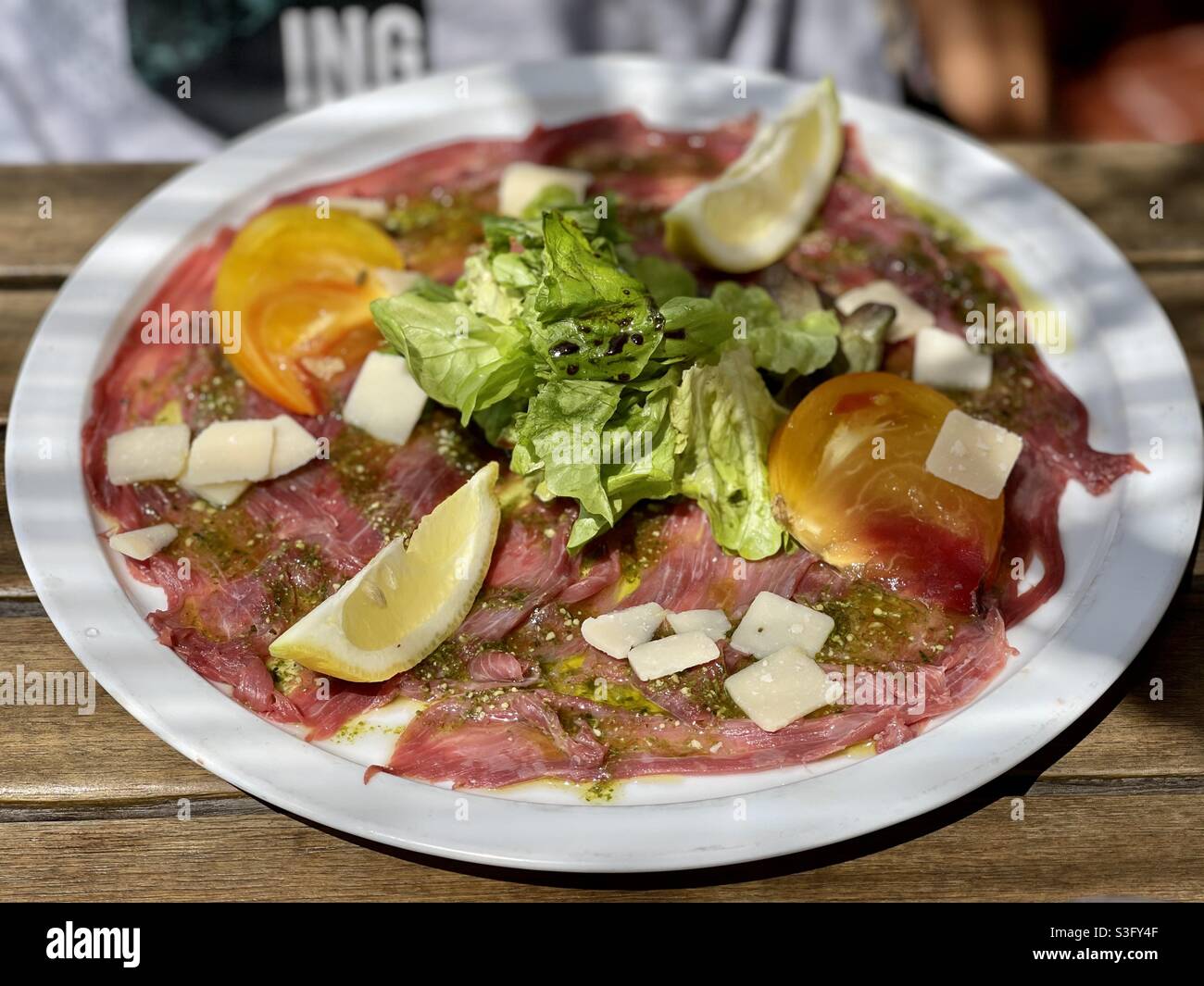 Red beef carpaccio with lemon, olive oil, yellow tomatoes, lettuce, balsamic vinegar, parmesan cheese and pesto on a restaurant plate on a wooden table in the south of France. - Smartphone Captured Stock Image