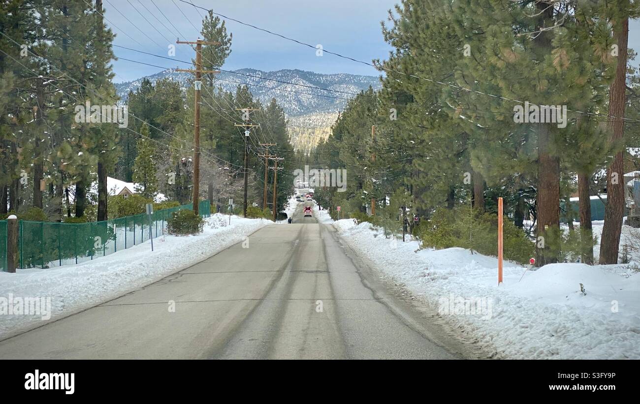 Road lined with ploughed snow and pine trees at Big Bear, California, on Christmas Eve, 2020, with overcast skies, telephone cables, and mountains in the background. Stock Photo