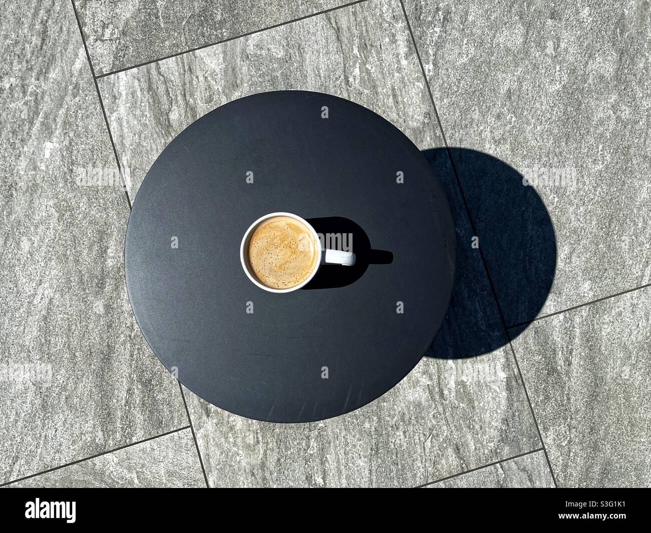 Overhead view of a cup of coffee on the plain dark top of a garden table standing on the grey porcelain paving slabs of patio Stock Photo