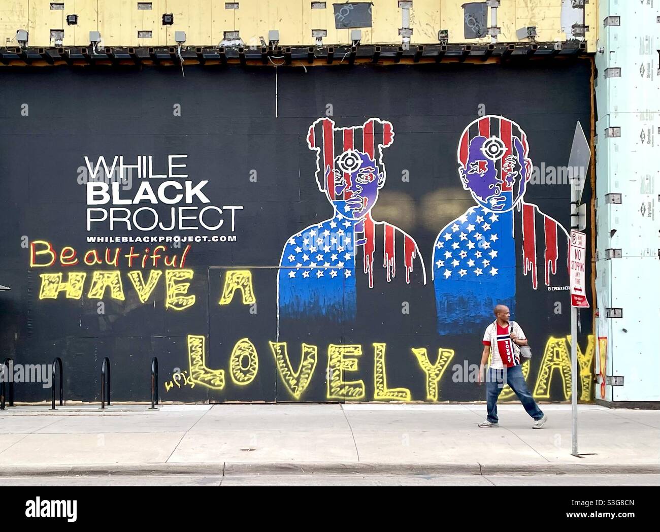 A black man wearing a Norway flag  t-shirt walking by street art advertising the While Black Project. African American man looking over his shoulder as if being followed. - Smartphone Captured Stock Image