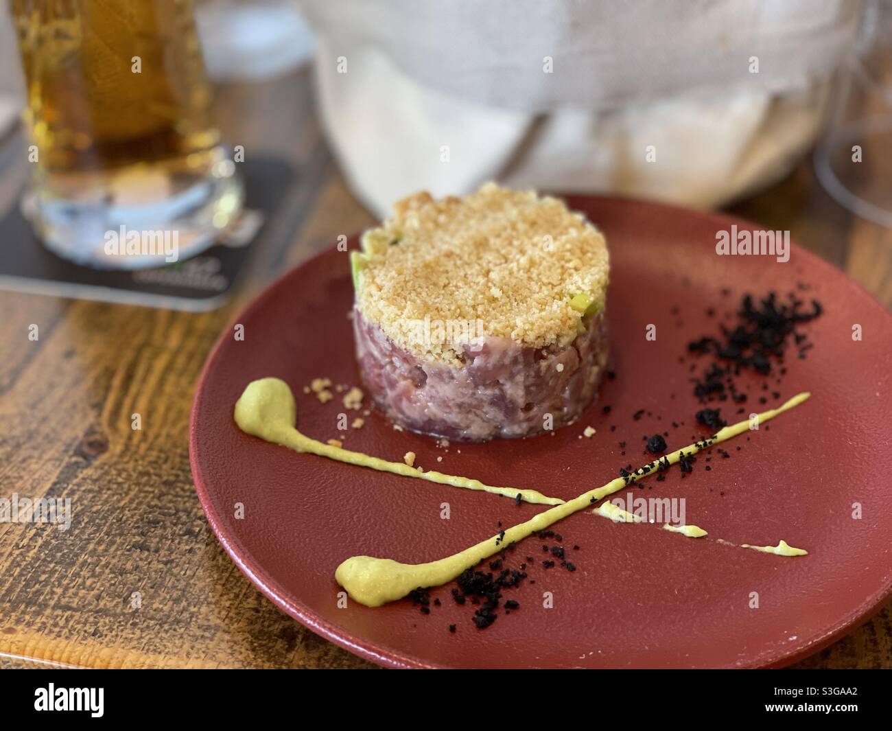 Red tuna tartare with radish sauce on a red restaurant plate on a wooden table, with a glass of beer and a basket of bread Stock Photo
