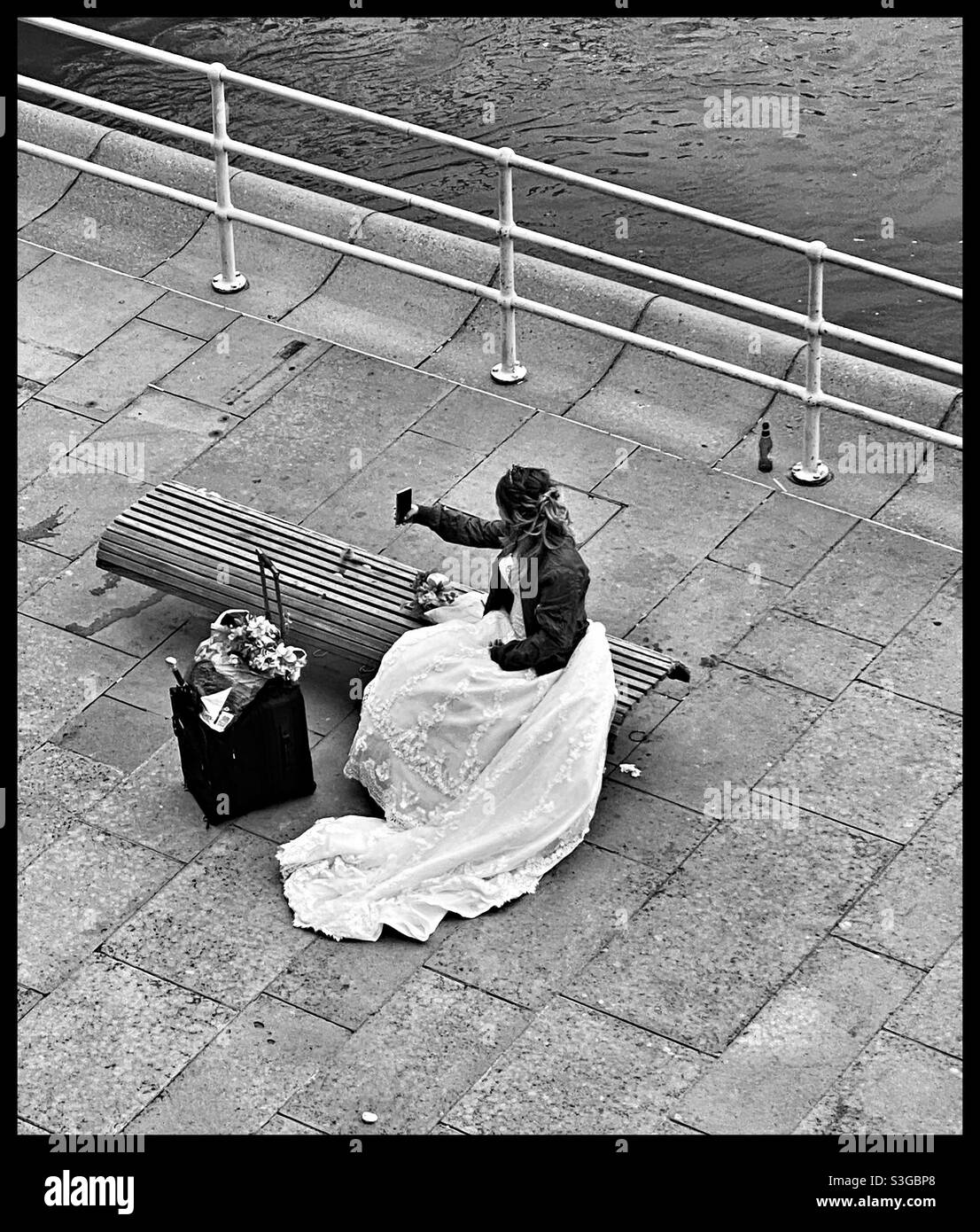 A bride in her wedding dress and a jacket, sitting alone on a bench by her suitcase, taking a selfie. Stock Photo