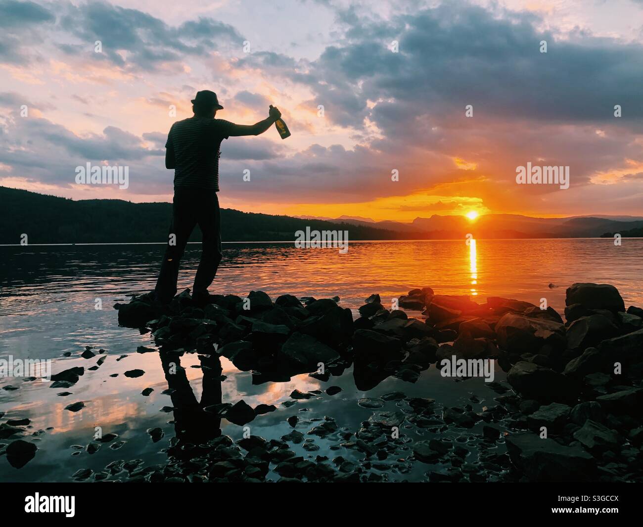 A man holding a bottle of Prosecco silhouette by the sunset at lake Windermere - Smartphone Captured Stock Image