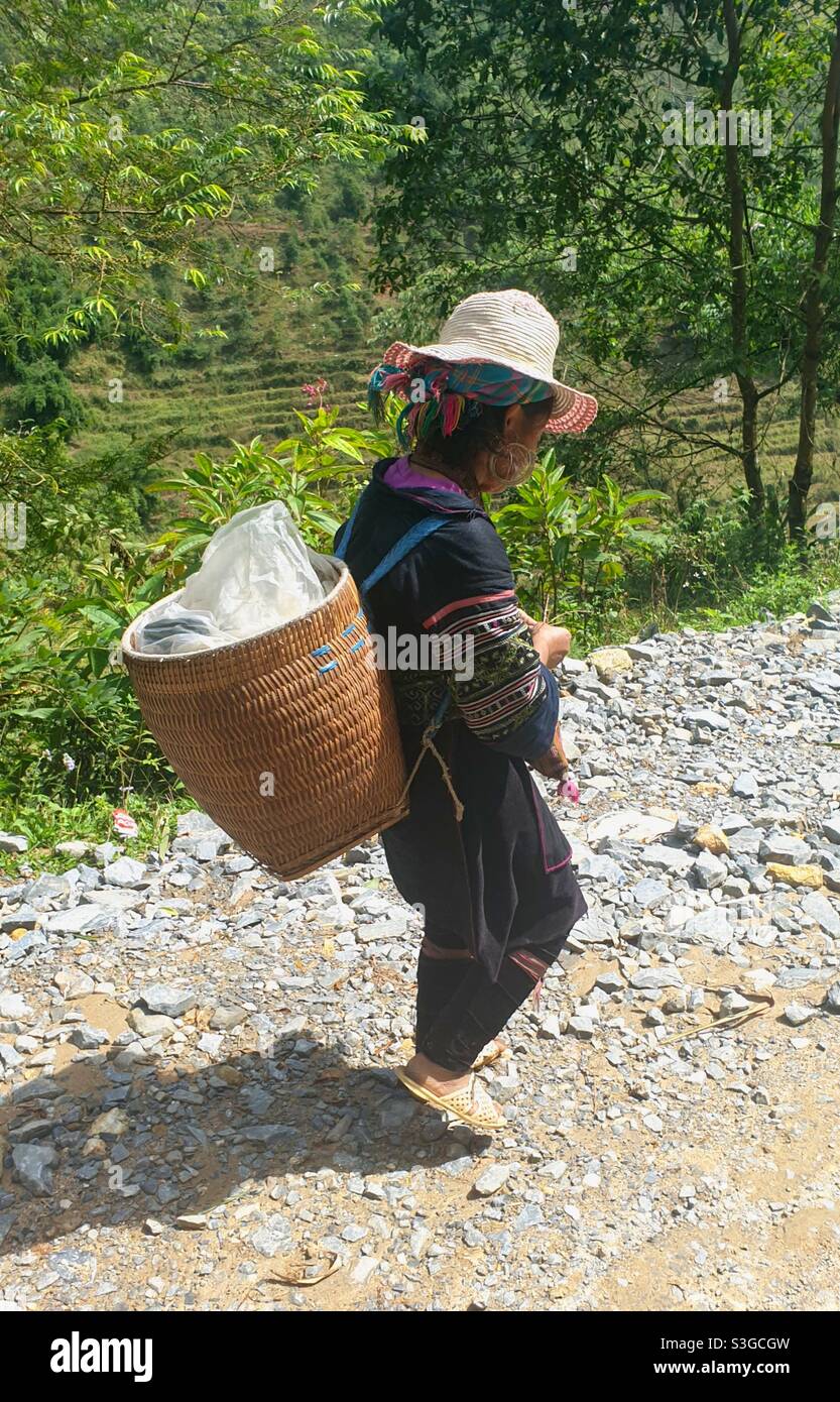 Black Hmong woman in traditional dress walking along the road with a basket of her embroidery items in Sapa, North Vietnam Stock Photo