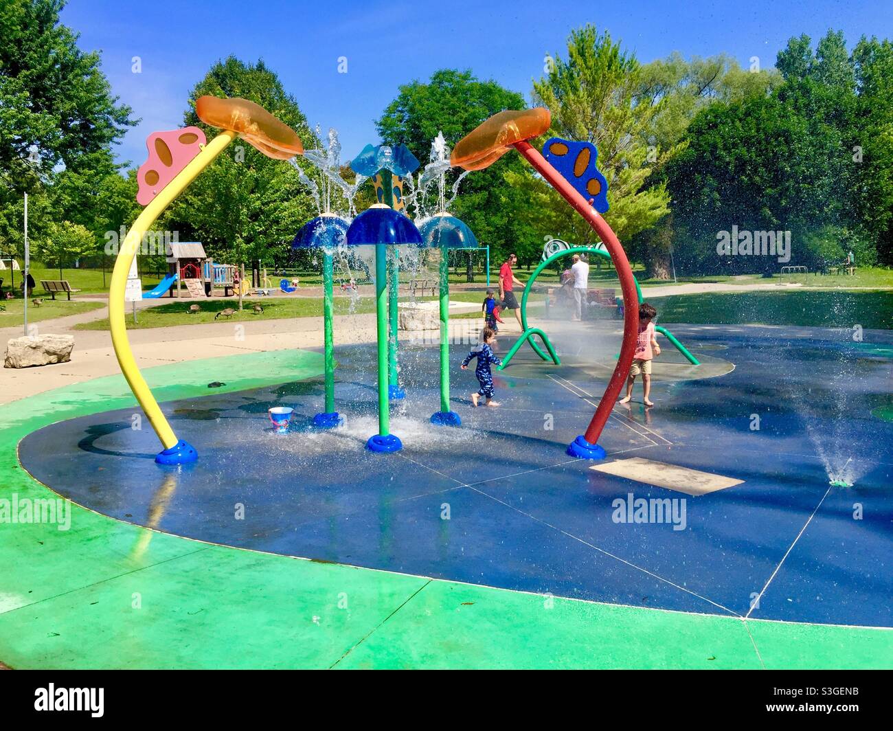 Outdoor fun on a modern splash pad with cascading water, sprays, and fountains, Ontario, Canada. Swings, slides, play equipment nearby. Stock Photo