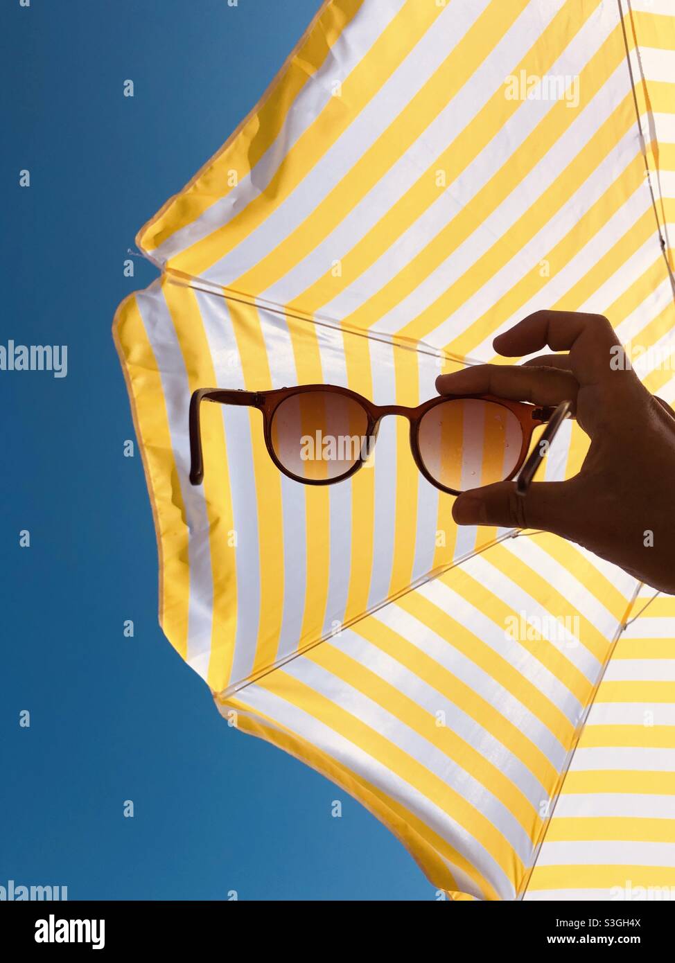 Man holding sunglasses against yellow and white striped beach umbrella - Smartphone Captured Stock Image