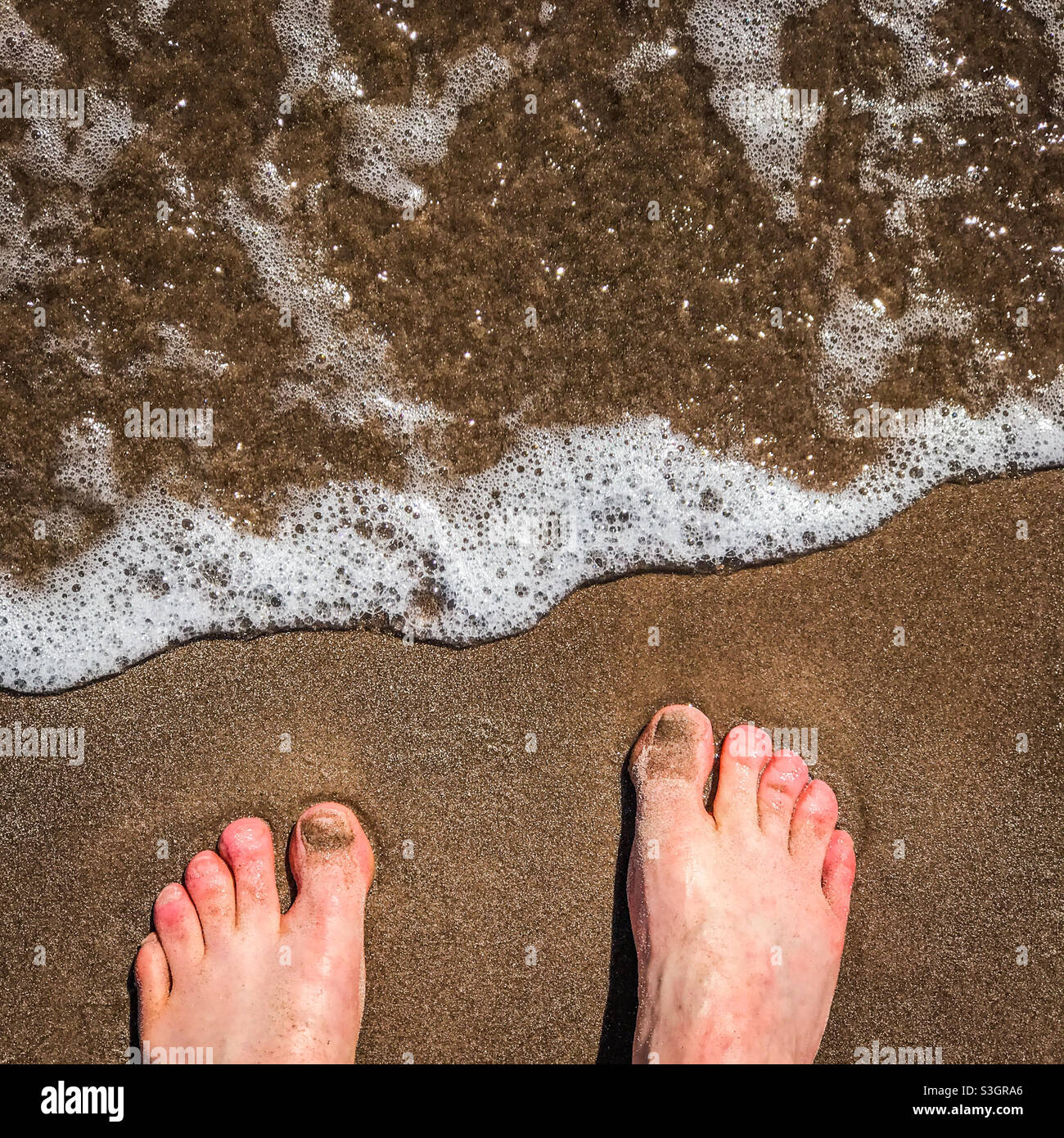Two feet on a sandy beach washed over by a wave - Smartphone Captured Stock Image