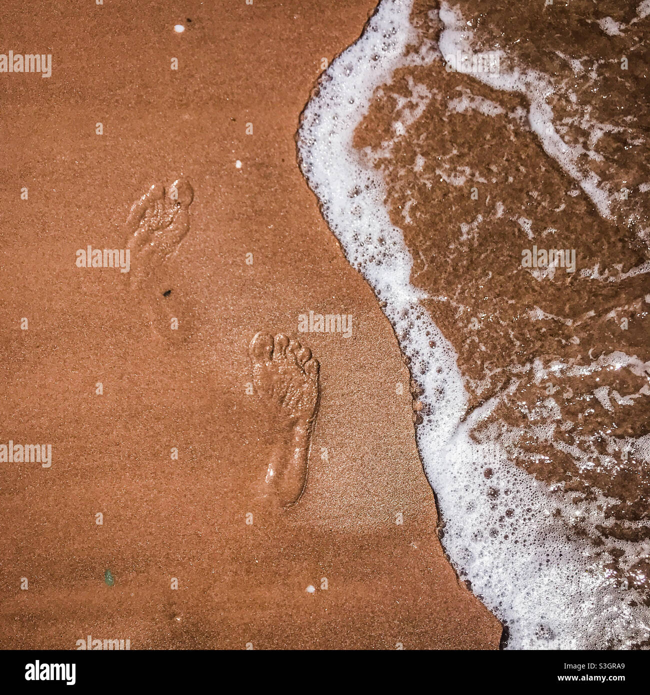 Two footprints in the sand on a beach being washed away by a wave - Smartphone Captured Stock Image