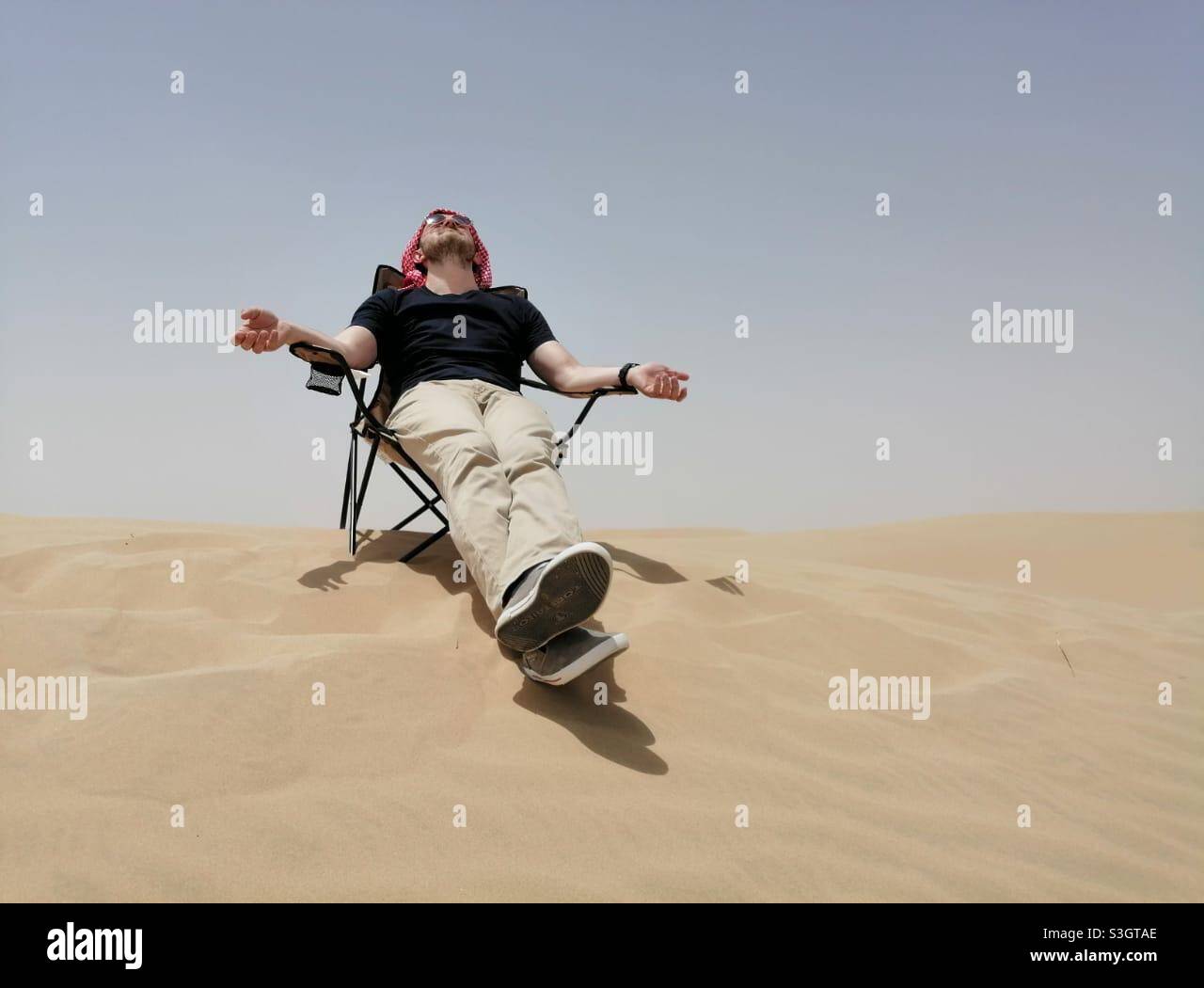 Tourist man enjoys the sun while sitting on a chair in desert sand dunes in Dubai, United Arab Emirates. Stock Photo