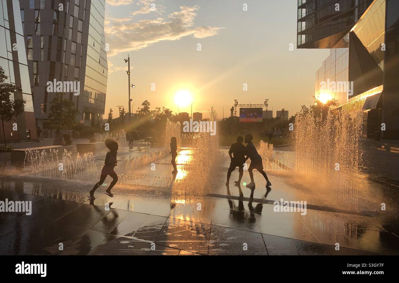 Four children playing in a fountain at sunset in Waterline Square Park, NYC. Friday, July 23, 2021. The Olympics is playing on a large screen in the background. Stock Photo