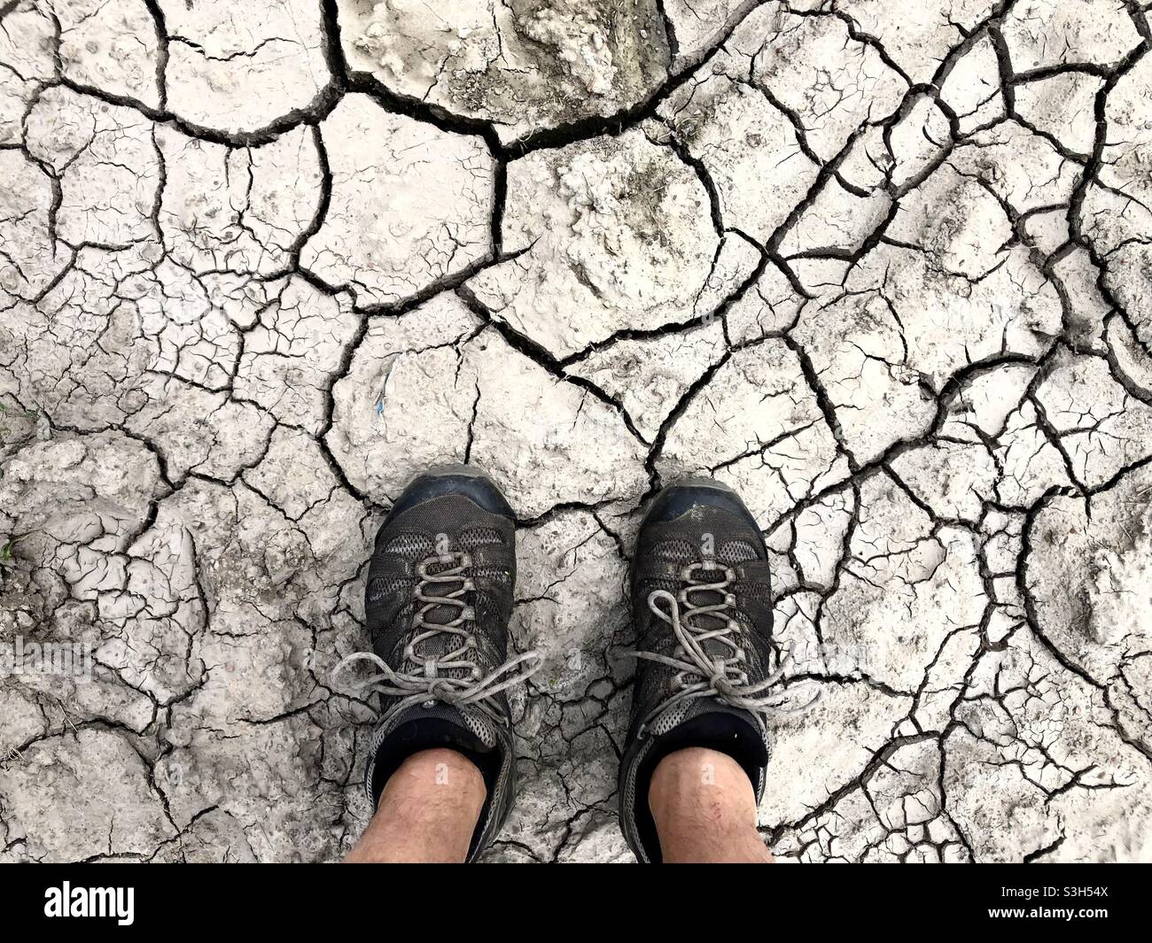 Looking down from above onto a pair of male hiking boots standing on arid and cracked earth during a dry spell of drought Stock Photo