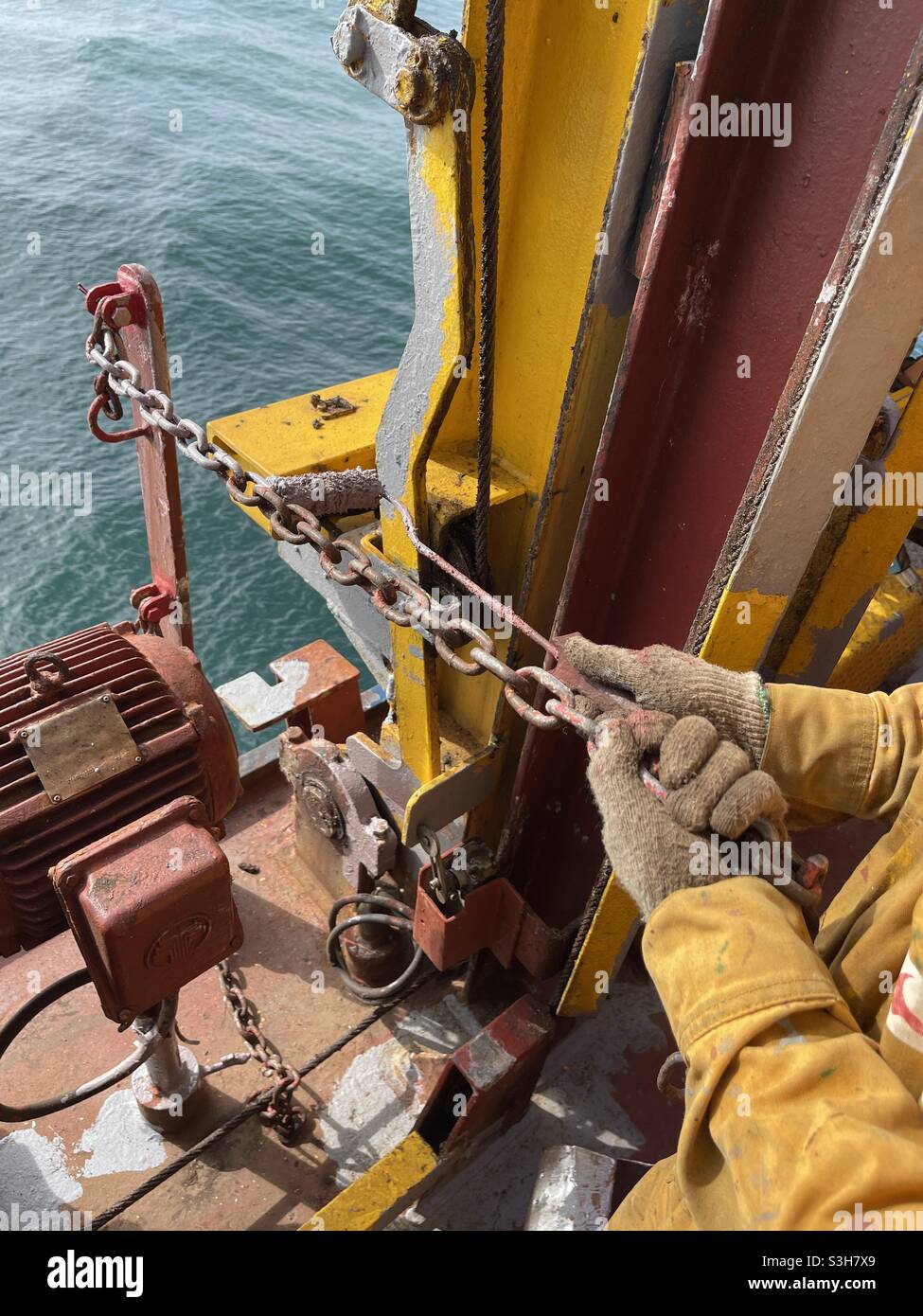 Crew member with cotton gloves of the merchant ship painting with primer paint of grey colour the chain of the pilot slant ladder using paint roller. Stock Photo