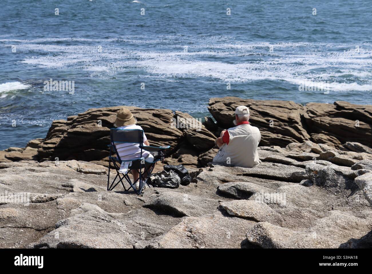 A couple, man and woman are sitting on the rocks and admire the waves splashing on the rocks and the ocean in Brittany - Smartphone Captured Stock Image