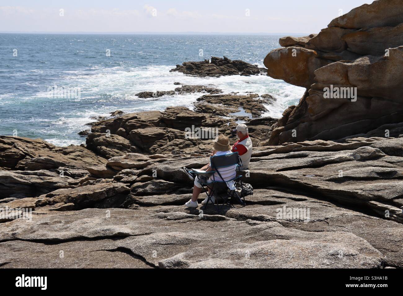 A couple, man and woman are sitting on the rocks and admire the waves splashing on the rocks and the ocean in Brittany  on July 2021 - Smartphone Captured Stock Image
