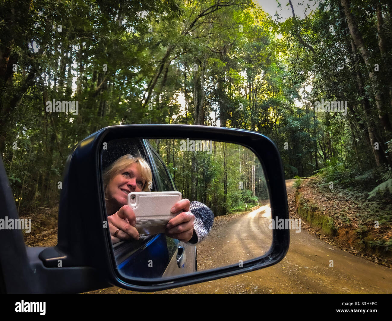 Beautiful blonde woman leaning out of a car window to take a photo of the Australian Bush in a national park along a winding dirt road, catching her reflection in the rear view mirror. Stock Photo