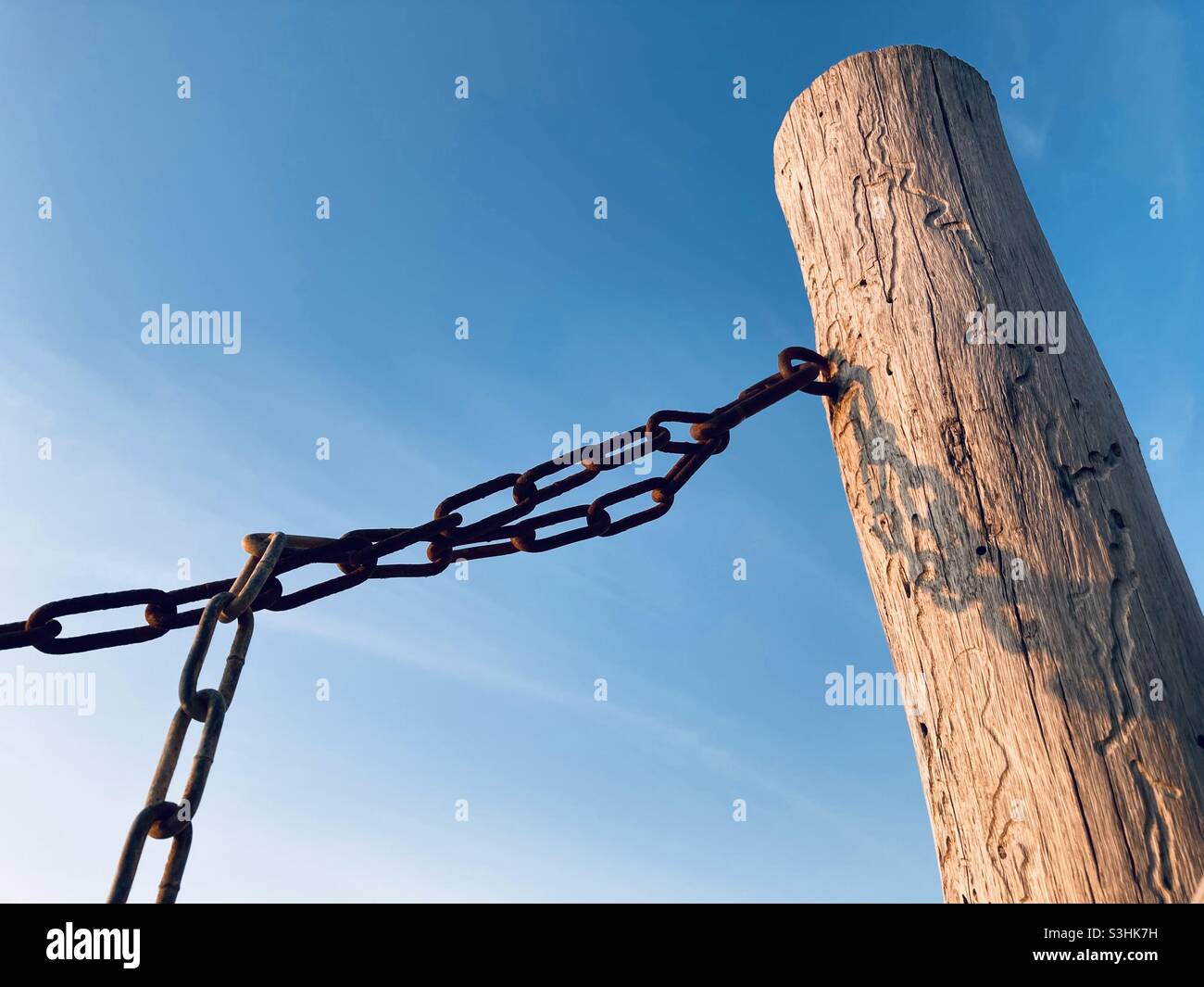 A rusty chain is attached to a wooden post against a blue sky in the evening sun - Smartphone Captured Stock Image
