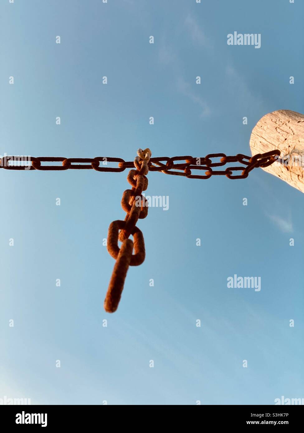 A rusty chain is attached to a wooden post against a blue sky in the evening sun - upview - Smartphone Captured Stock Image