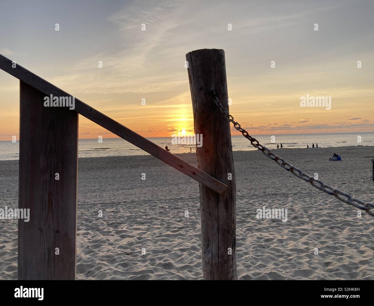 A rusty chain is attached to a wooden pole on the beach, in front of the sunset in the background - Smartphone Captured Stock Image