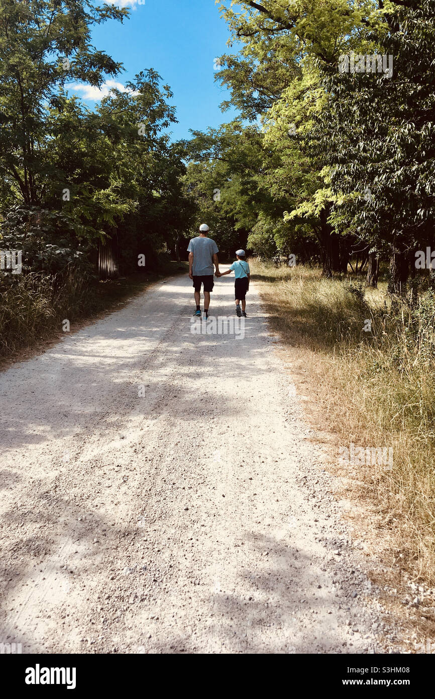 Father and son walking away on dirt road lined with trees in summer - Smartphone Captured Stock Image